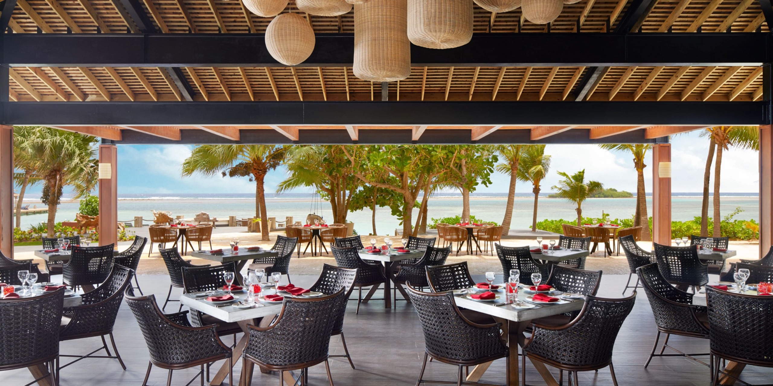 The Zoetry Montego Bay offers a charming outdoor tropical dining experience, featuring wicker chairs and tables elegantly adorned with red napkins. Enjoy the scenic view of palm trees and the ocean while hanging lanterns enhance the ambiance of the wooden ceiling.