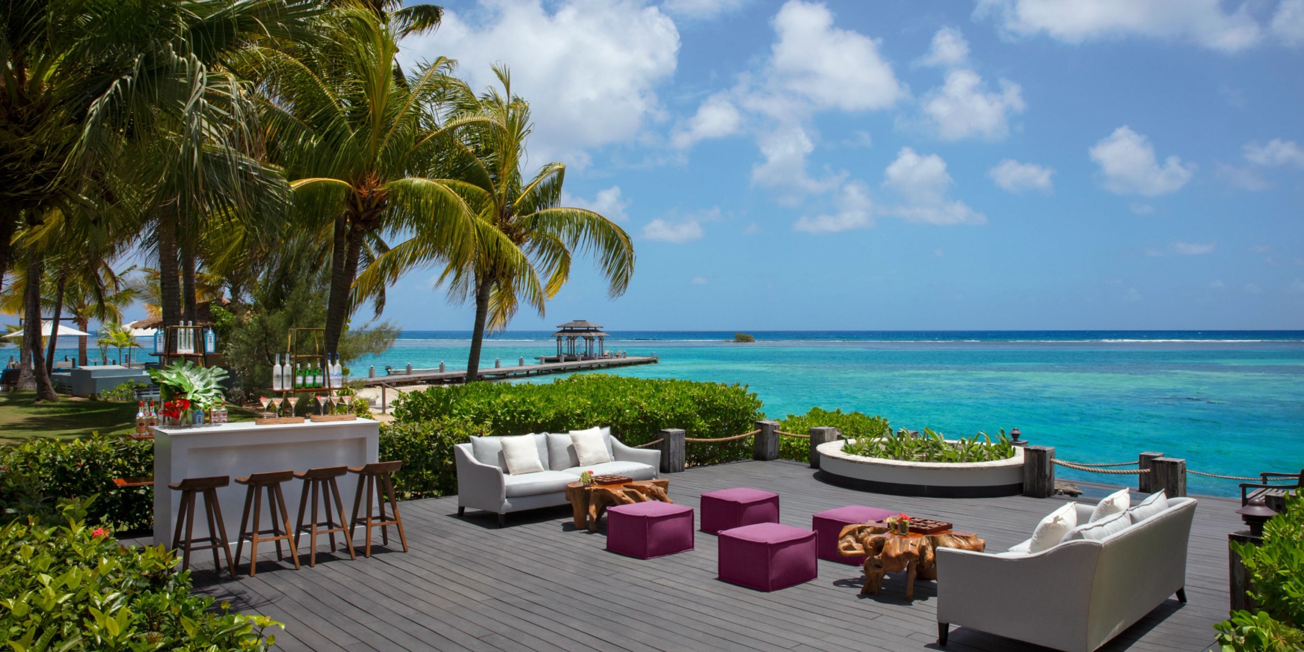 Relax in the Zoetry Montego Bay's tropical outdoor lounge, featuring sofas and stools on a deck with stunning views of the ocean and palm trees under a clear blue sky.