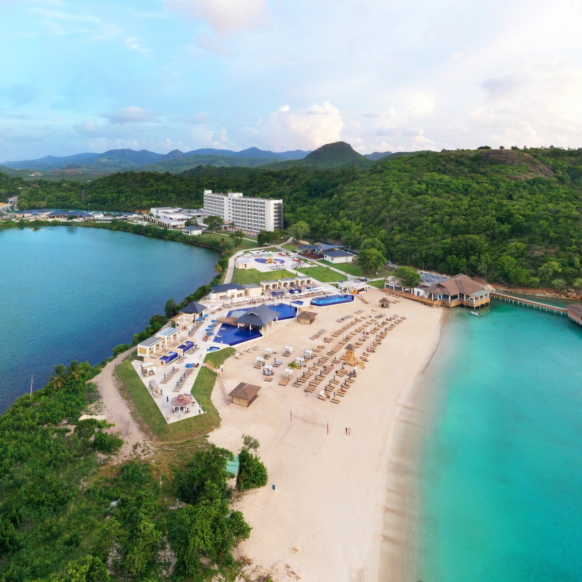 Aerial view of Royalton Antigua with sandy beach, blue pools, and surrounded by lush green landscape and calm blue waters.