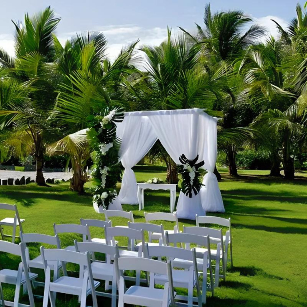 Destination wedding setup with white chairs and a floral-decorated arch, surrounded by lush green palm trees under clear skies.