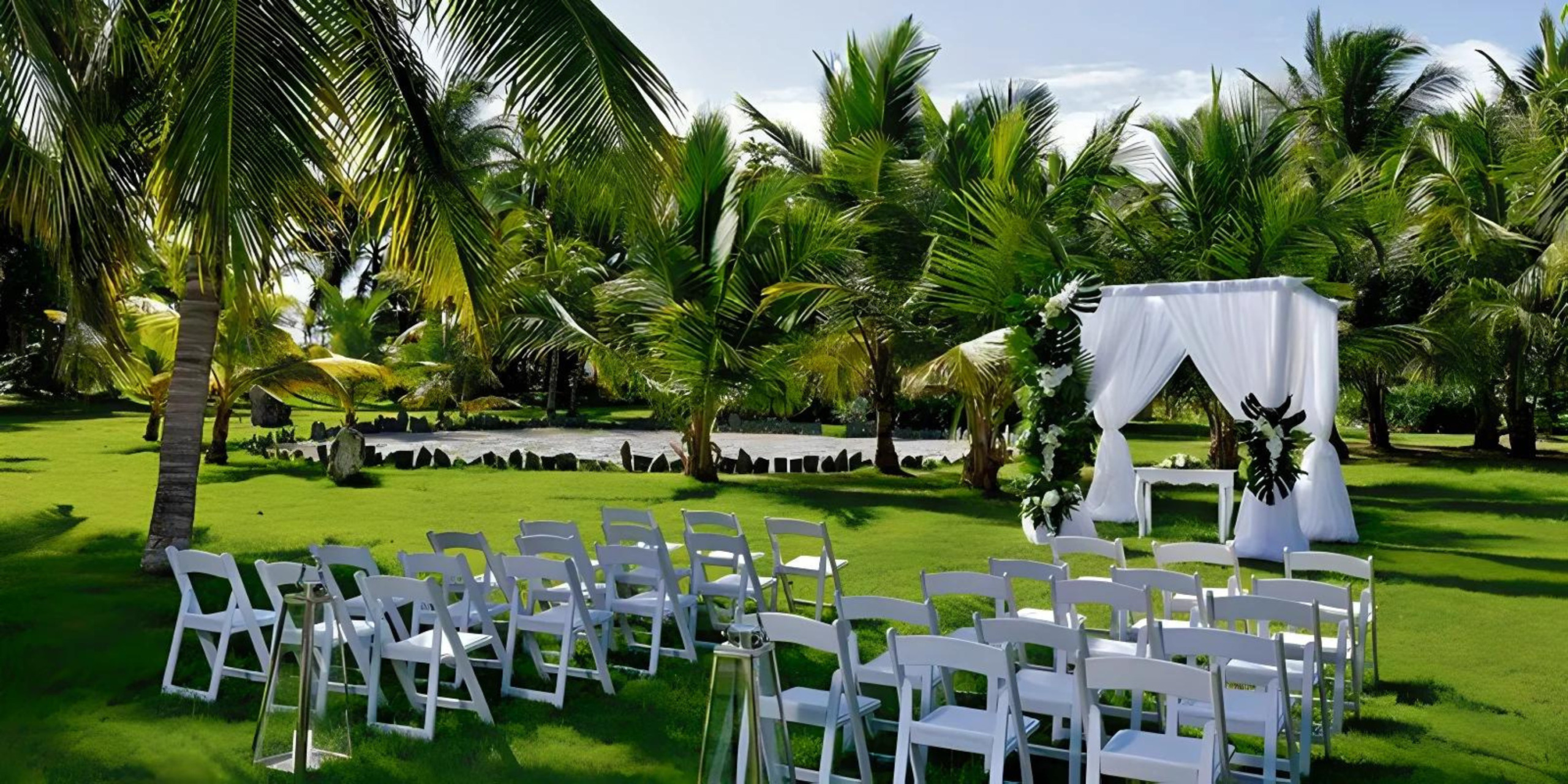 Destination wedding setup with rows of white chairs facing an altar under a canopy, surrounded by lush palm trees on a manicured lawn.
