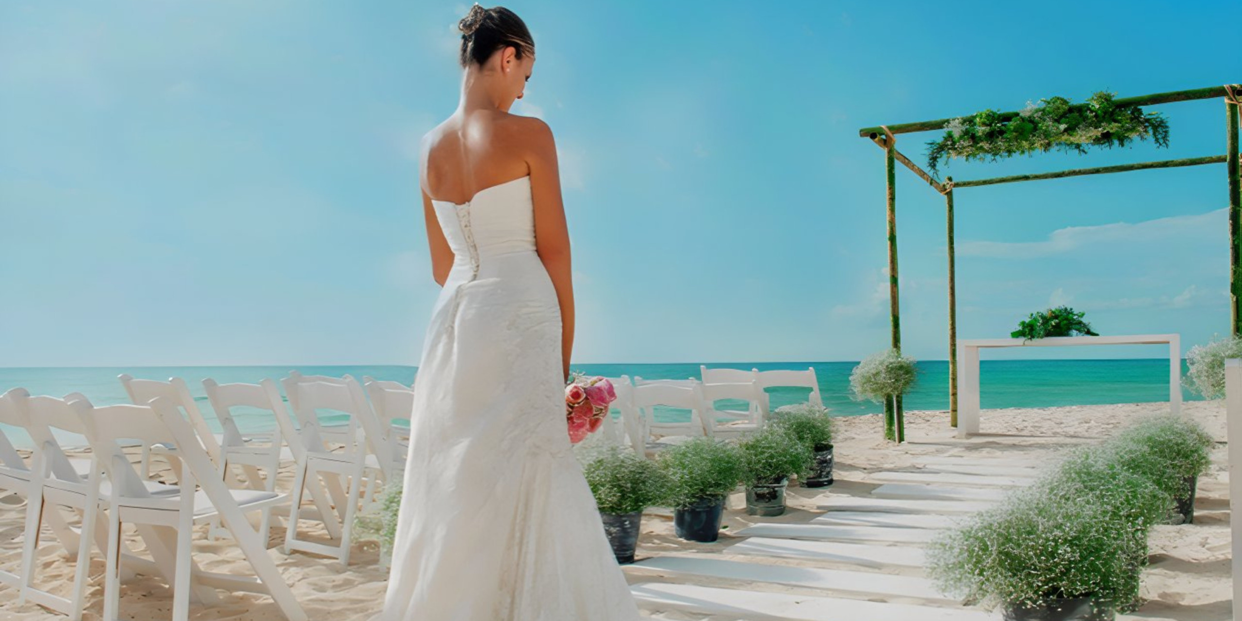 A bride in a white dress holding a bouquet walks along a sandy aisle between rows of white chairs at a destination beach wedding setting.