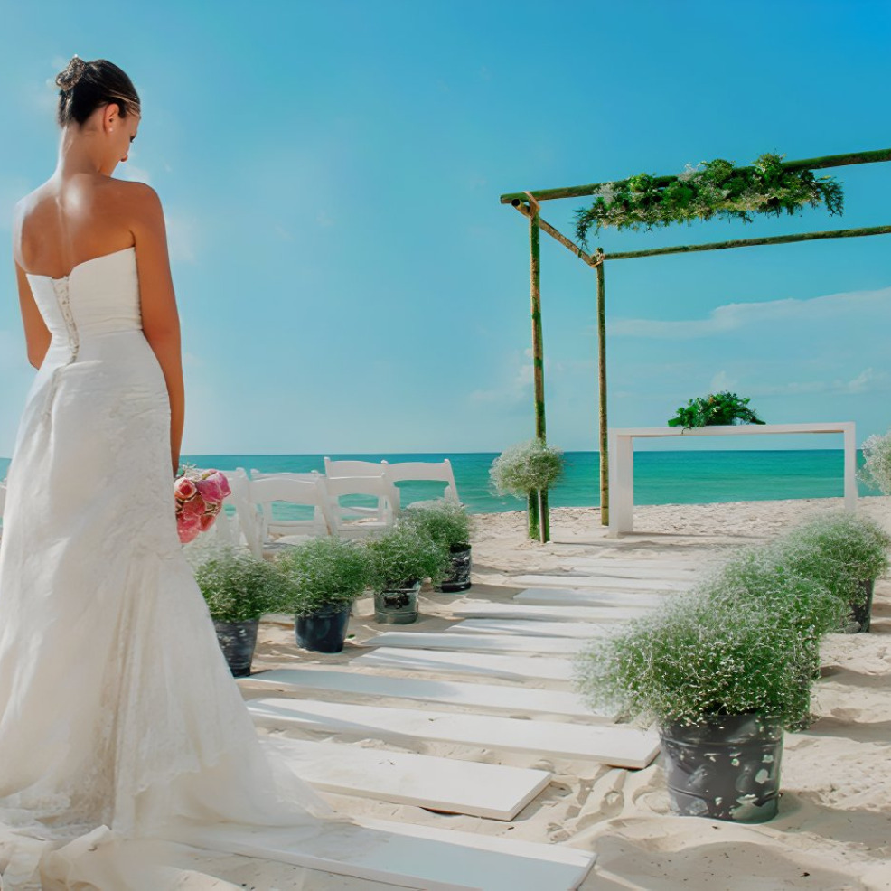 A bride in a white dress holding a bouquet stands by a destination wedding setup with white chairs and a floral arch, overlooking the ocean.