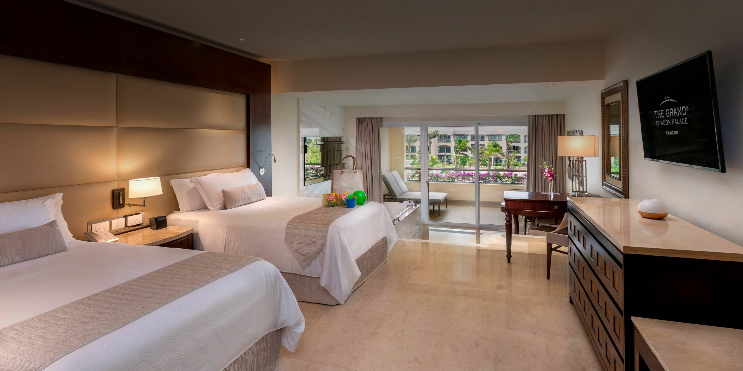 A hotel room with two double beds, a dresser, a wall-mounted TV, and a spacious seating area with a sofa and a window overlooking a balcony with a garden view at Moon Palace The Grand.