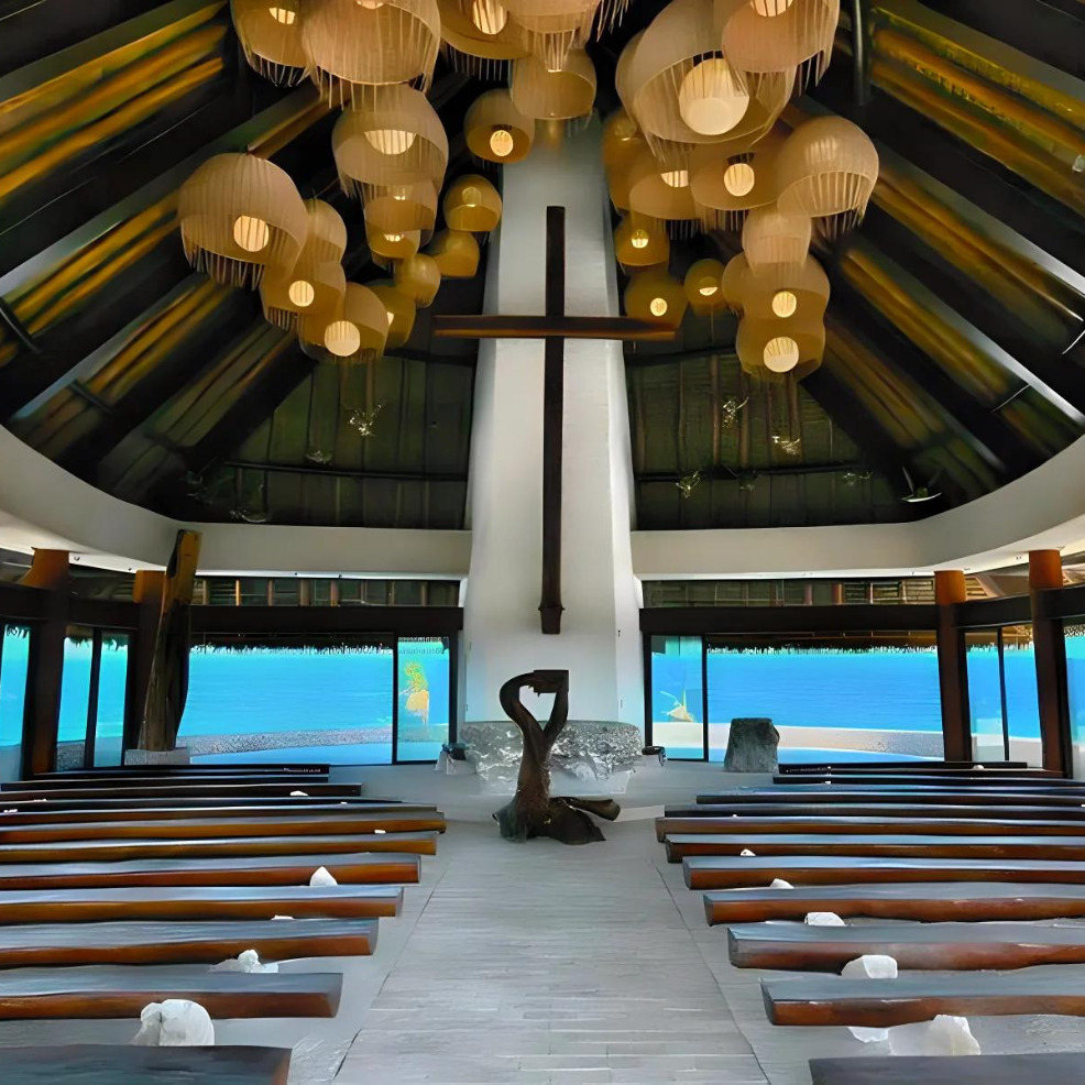 Interior of a modern chapel with wooden pews, a central cross, and hanging lanterns, fronting a large window overlooking the ocean, perfect for a destination wedding.