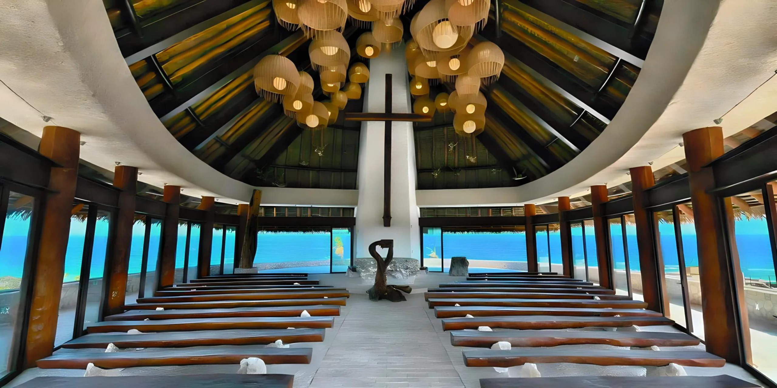 Interior of a modern chapel with symmetrical wooden beams, pendant lights, and benches facing an ocean view framed by large windows, perfect for a destination wedding.
