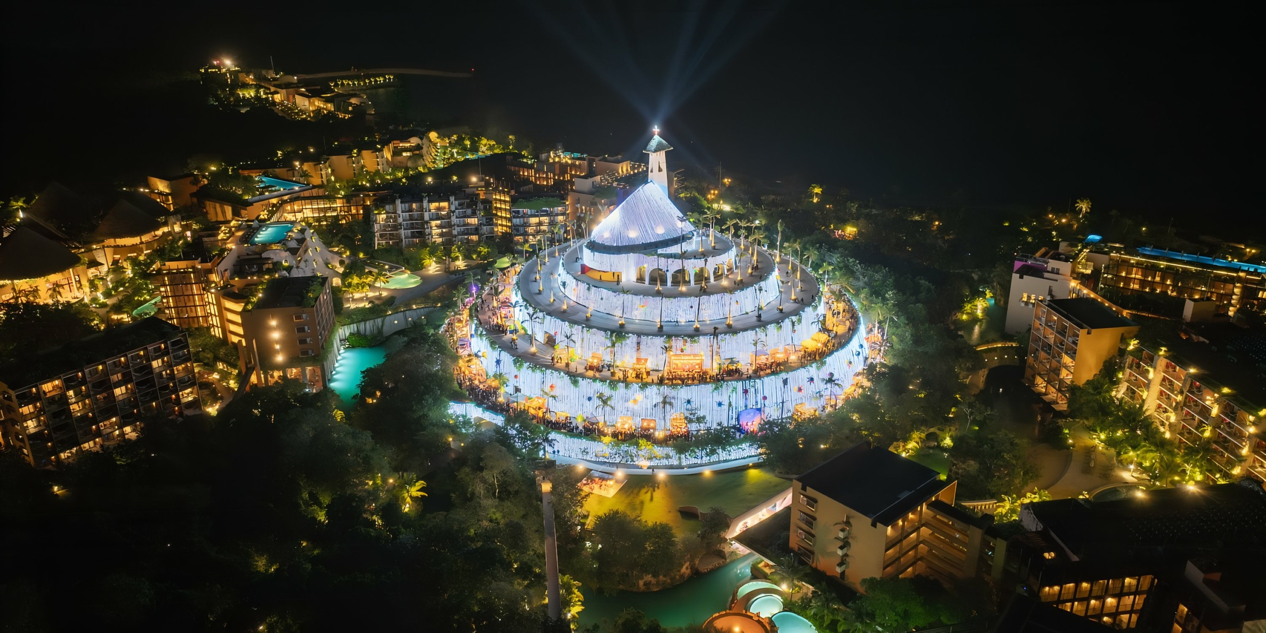 Aerial night view of Hotel Xcaret, a brightly lit, crowded resort complex with a large domed building at the center surrounded by lush greenery and buildings.
