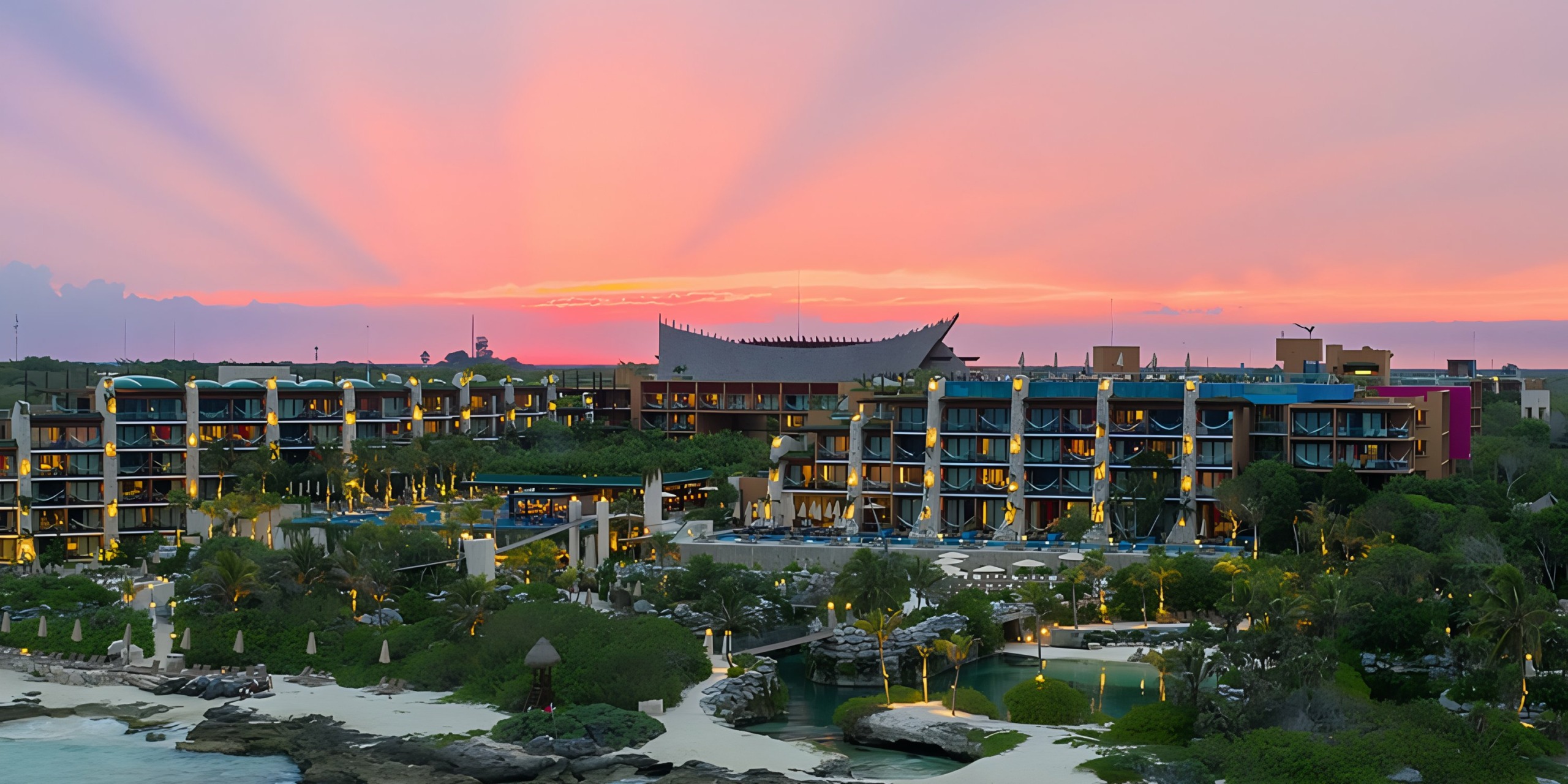 A luxury resort (Hotel Xcaret) with colorful buildings overlooking a tropical coastline at sunset, with visible pools and lush greenery under a vibrant pink and orange sky.