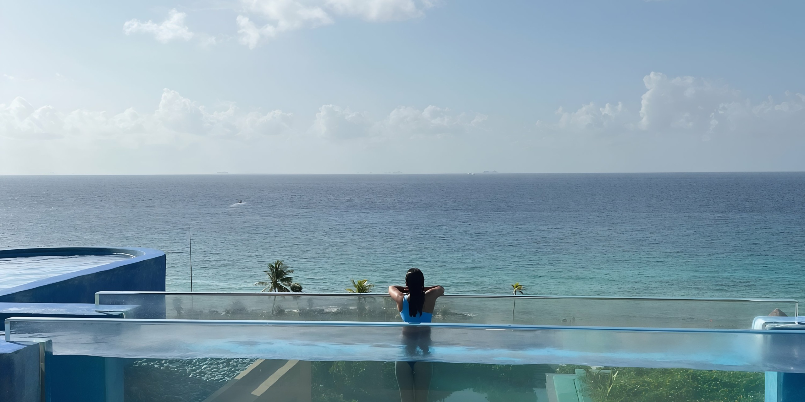 A person sits by an infinity pool overlooking the ocean at Hotel Xcaret, with a clear blue sky above and ships in the distance.