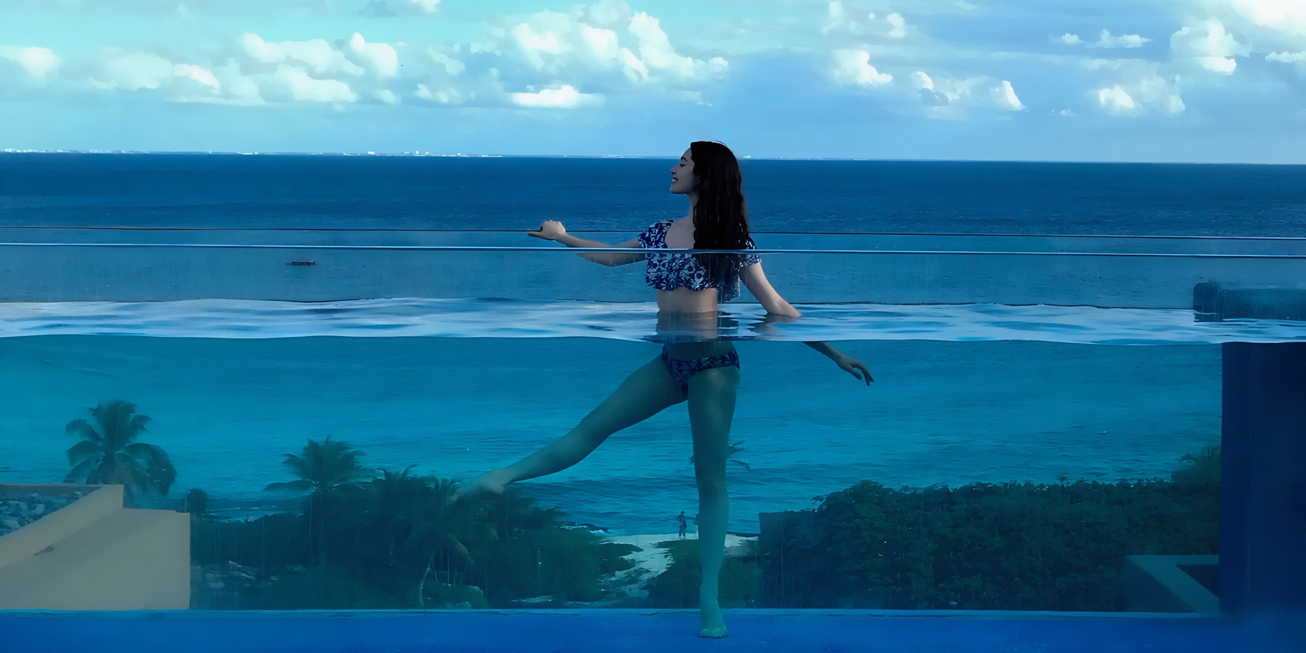 A woman balances on one leg on a Hotel Xcaret balcony railing overlooking a turquoise sea under a cloudy sky.