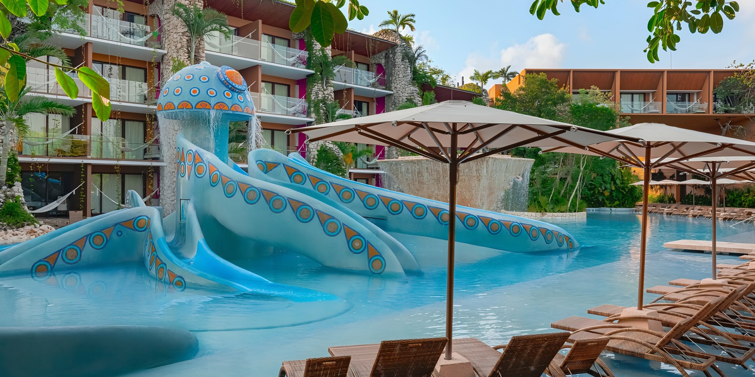 Resort pool area with Hotel Xcaret-themed slide, sun loungers under umbrellas, and surrounding hotel buildings.