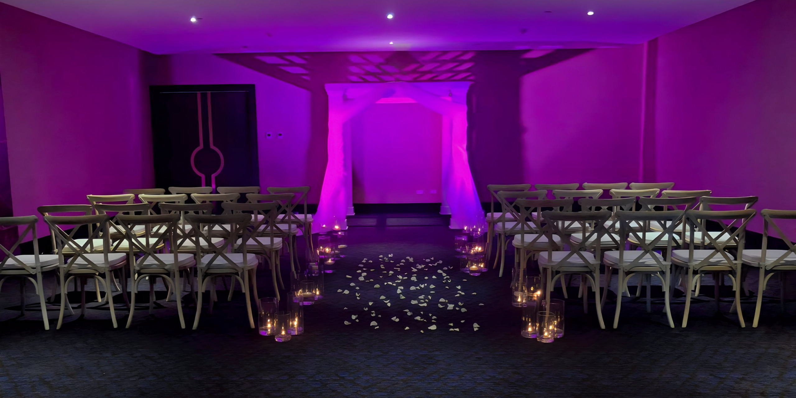 Elegant destination wedding aisle with white chairs and rose petals, illuminated by purple lighting in a modern venue.