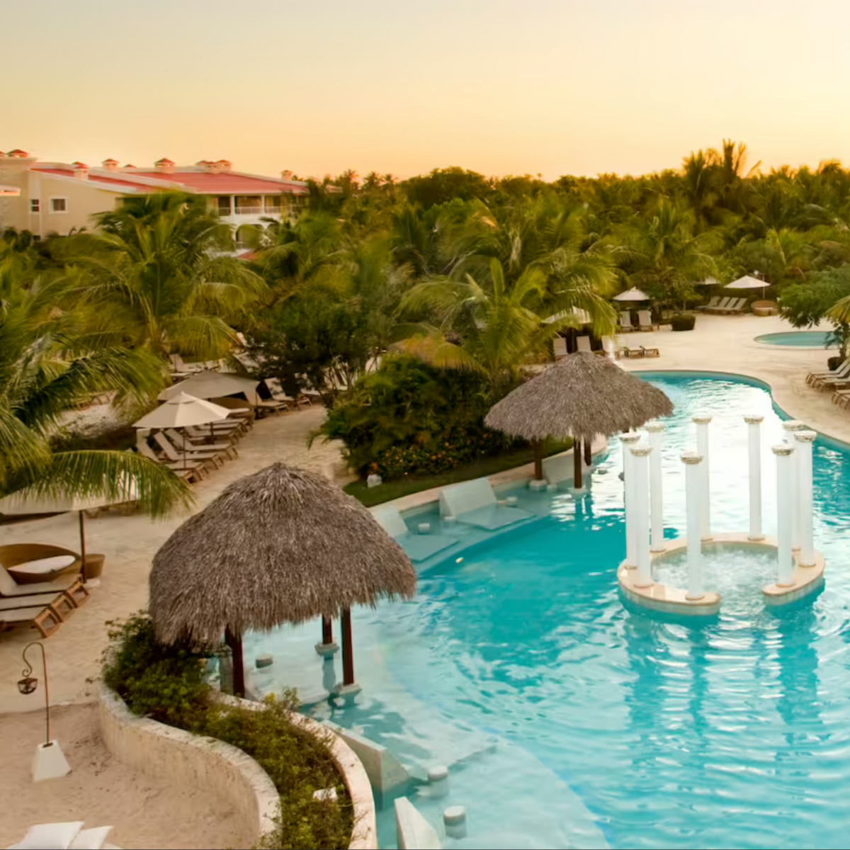 A luxurious Melia Caribe Beach resort pool at sunset, perfect for destination weddings, featuring thatched-roof cabanas and elegant white pillars surrounded by palm trees.