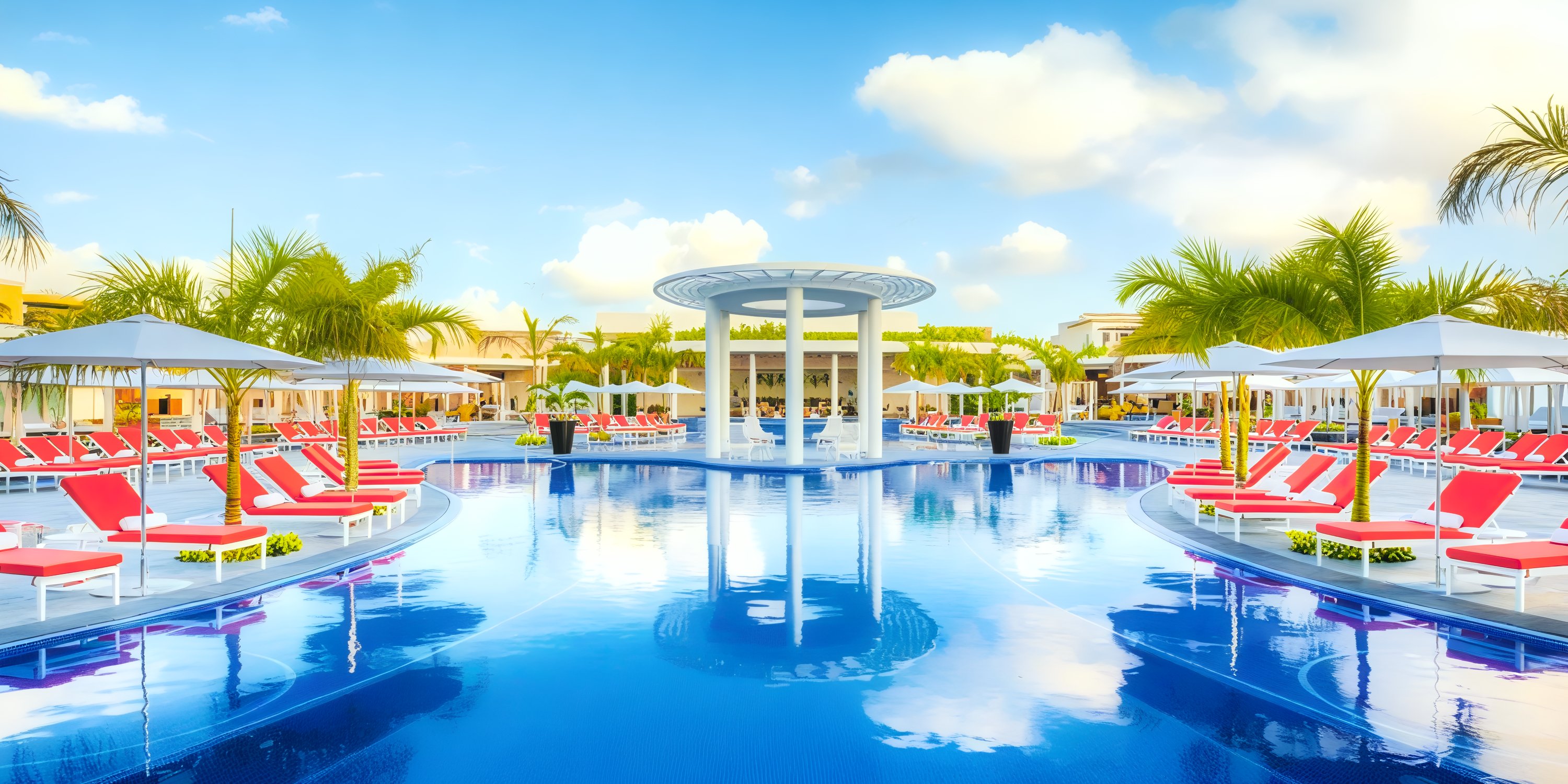 Moon Palace The Grand: A stunning pool area adorned with red lounge chairs and umbrellas, set against a backdrop of a cloudy blue sky—perfect for a destination wedding.