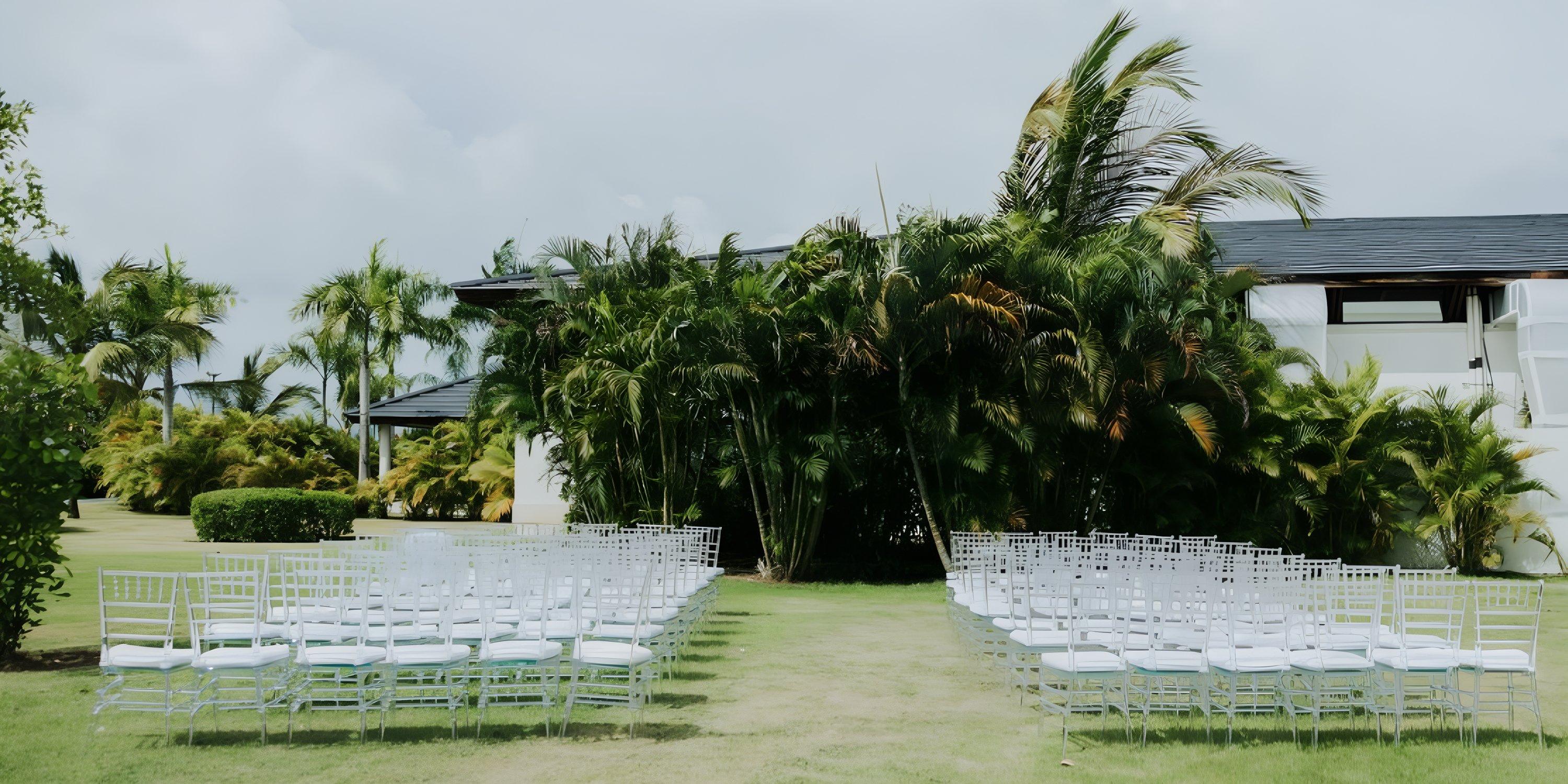 White wedding chairs set up outdoors at Ocean El Faro, facing palm trees and a building under cloudy skies.