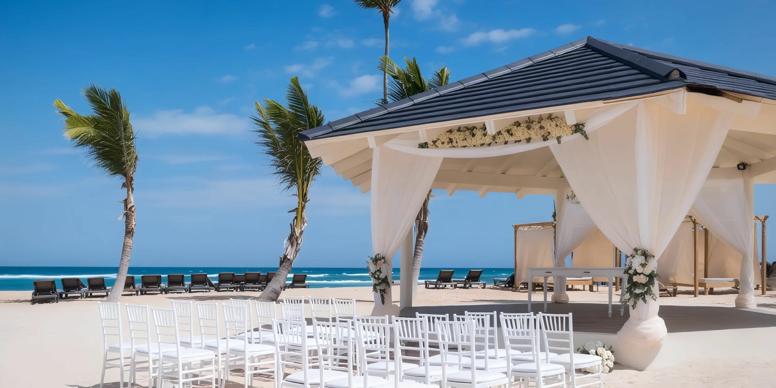 A beach wedding setup with white chairs facing an altar under a gazebo, decorated with flowers, against a backdrop of palm trees, sand, and ocean.