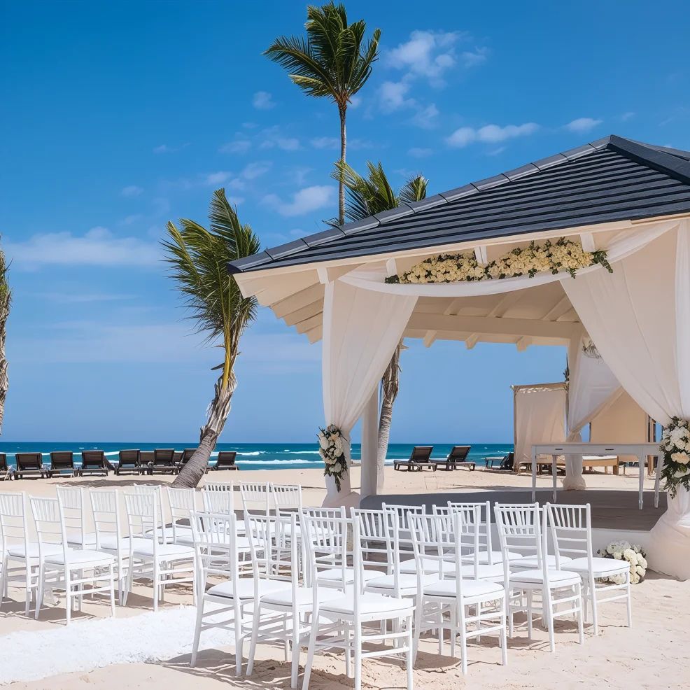 Beachfront wedding setup with white chairs facing a gazebo adorned with flowers. Palm trees and ocean in the background under a clear blue sky.