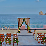 Destination wedding setup on a terrace overlooking the sea, with a floral arch and rows of wooden chairs.