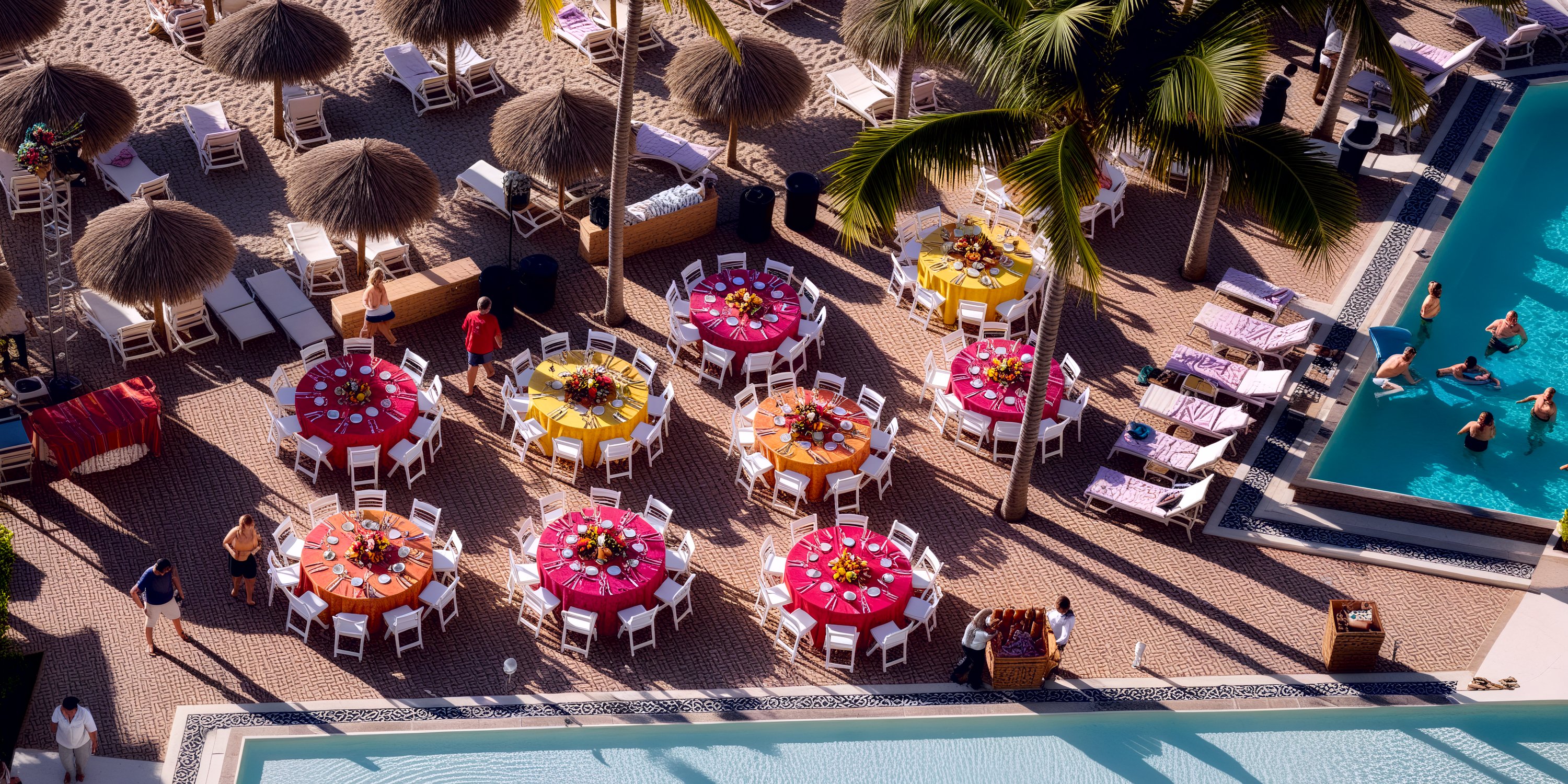 Vibrant tables are elegantly arranged by the poolside, adorned with umbrellas and framed by swaying palm trees, setting the scene for a picturesque destination wedding. Guests mingle joyfully around and in the pool, adding to the celebratory atmosphere.
