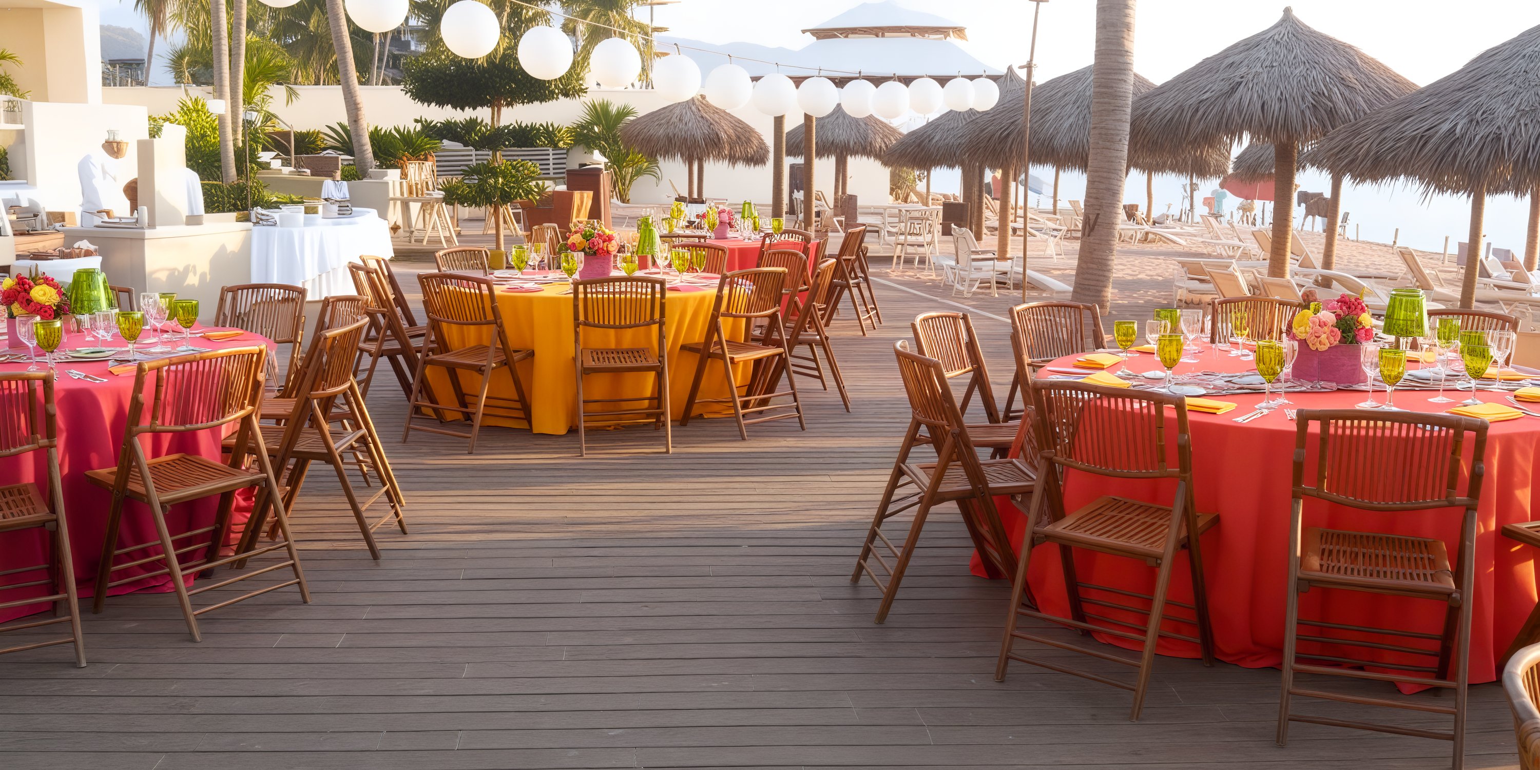 A destination wedding dining setup featuring vibrant tables and chairs nestled beneath palm-thatched umbrellas.