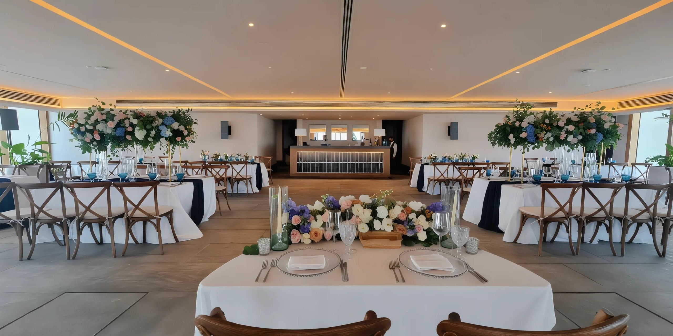 Elegant indoor venue with neatly arranged tables adorned with floral centerpieces, white tablecloths, and formal place settings, facing a well-lit bar area in the background.