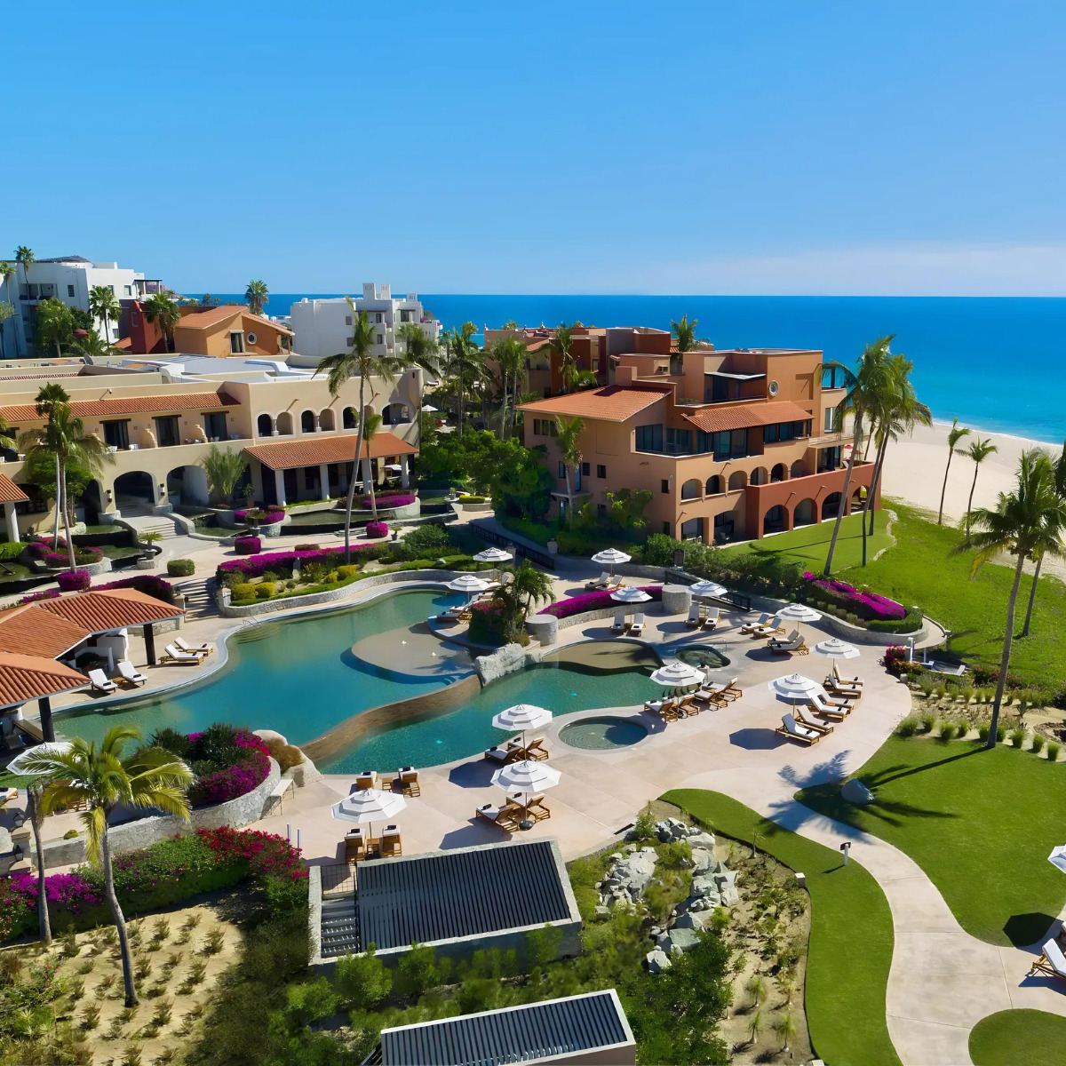 Aerial view of a luxury Zoetry Casa Del Mar beachfront resort with a large swimming pool, palm trees, and colorful gardens in a tropical setting.
