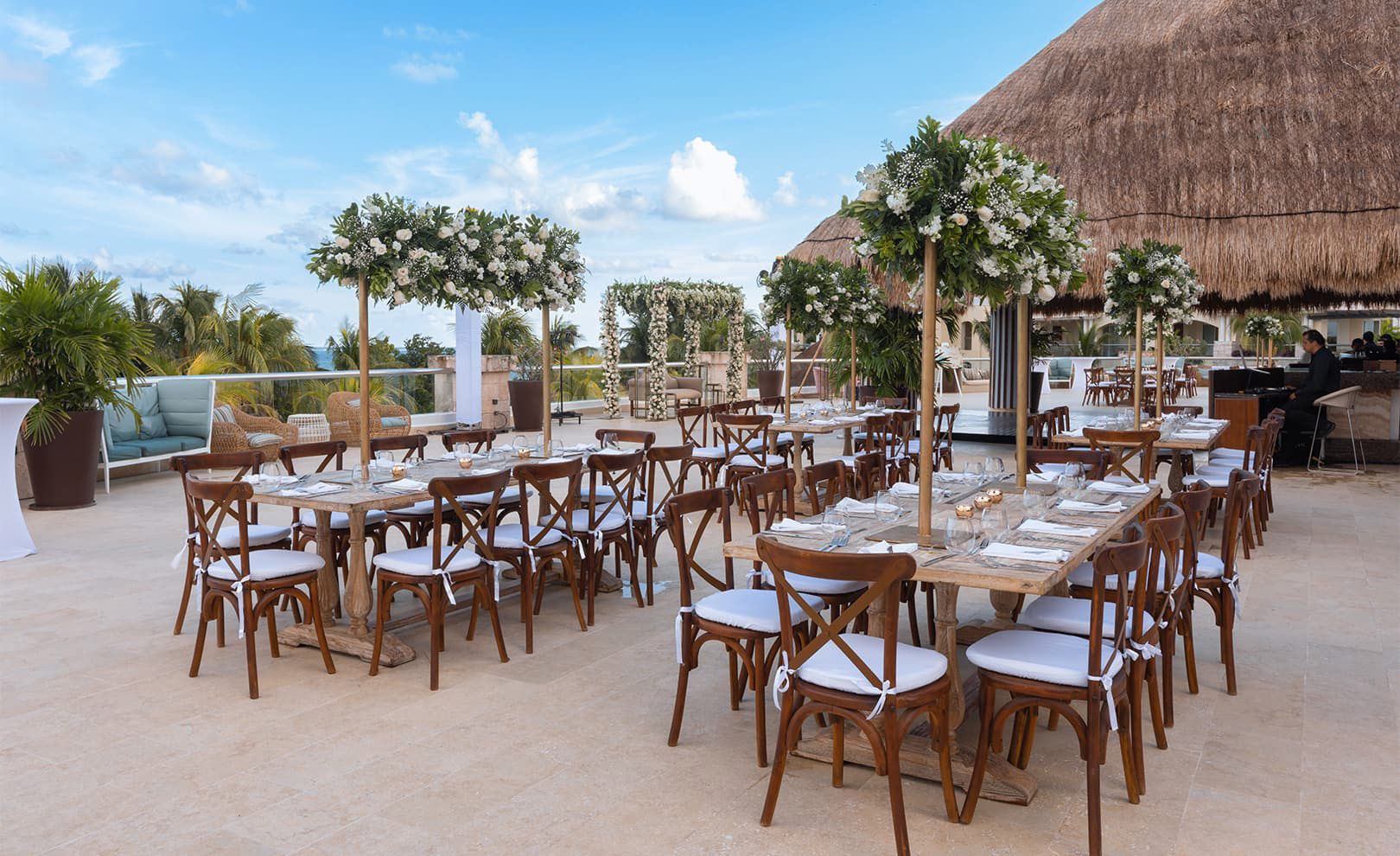 An outdoor reception area with wooden tables and chairs set up, featuring floral centerpieces and greenery. Moon Palace The Grand is in the background, under a clear blue sky.