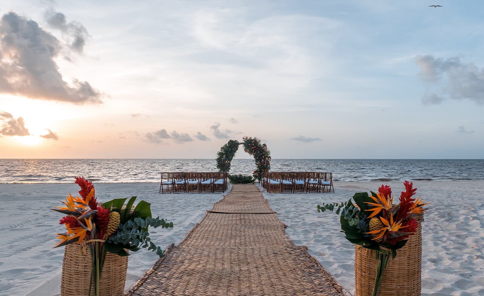 A Moon Palace The Grand setup with an aisle lined with woven mats, leading to a floral arch and rows of chairs facing the ocean at sunset.