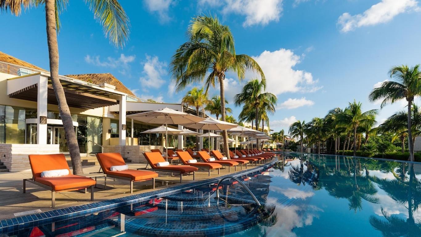 Moon Palace The Grand with orange lounge chairs, sun umbrellas, and palm trees. Modern buildings with open terraces in the background under a clear, blue sky.