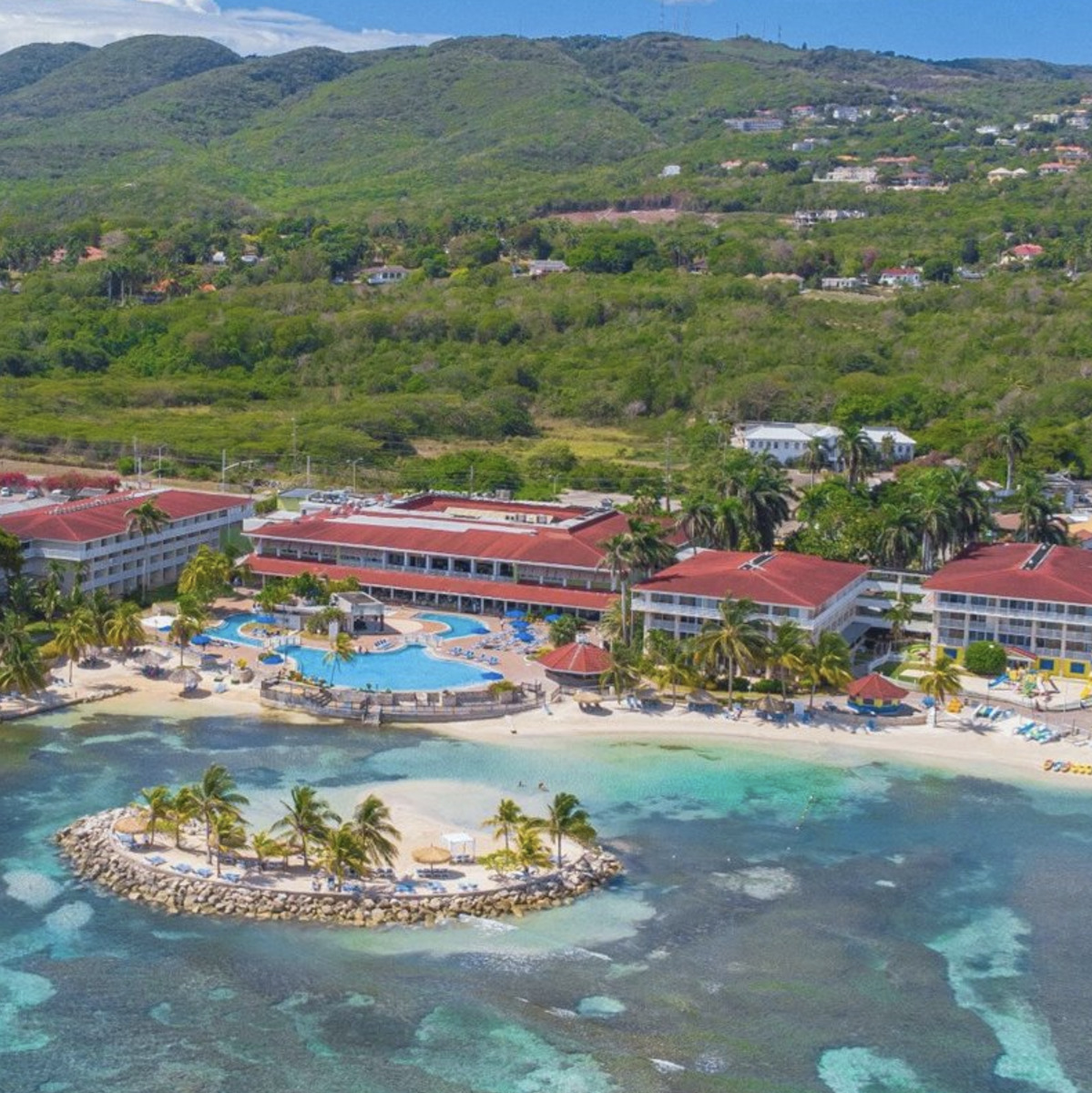 Aerial view of Holiday Inn Montego Bay, a tropical destination wedding resort with a large pool and surrounding villas, set against a backdrop of lush green hills.