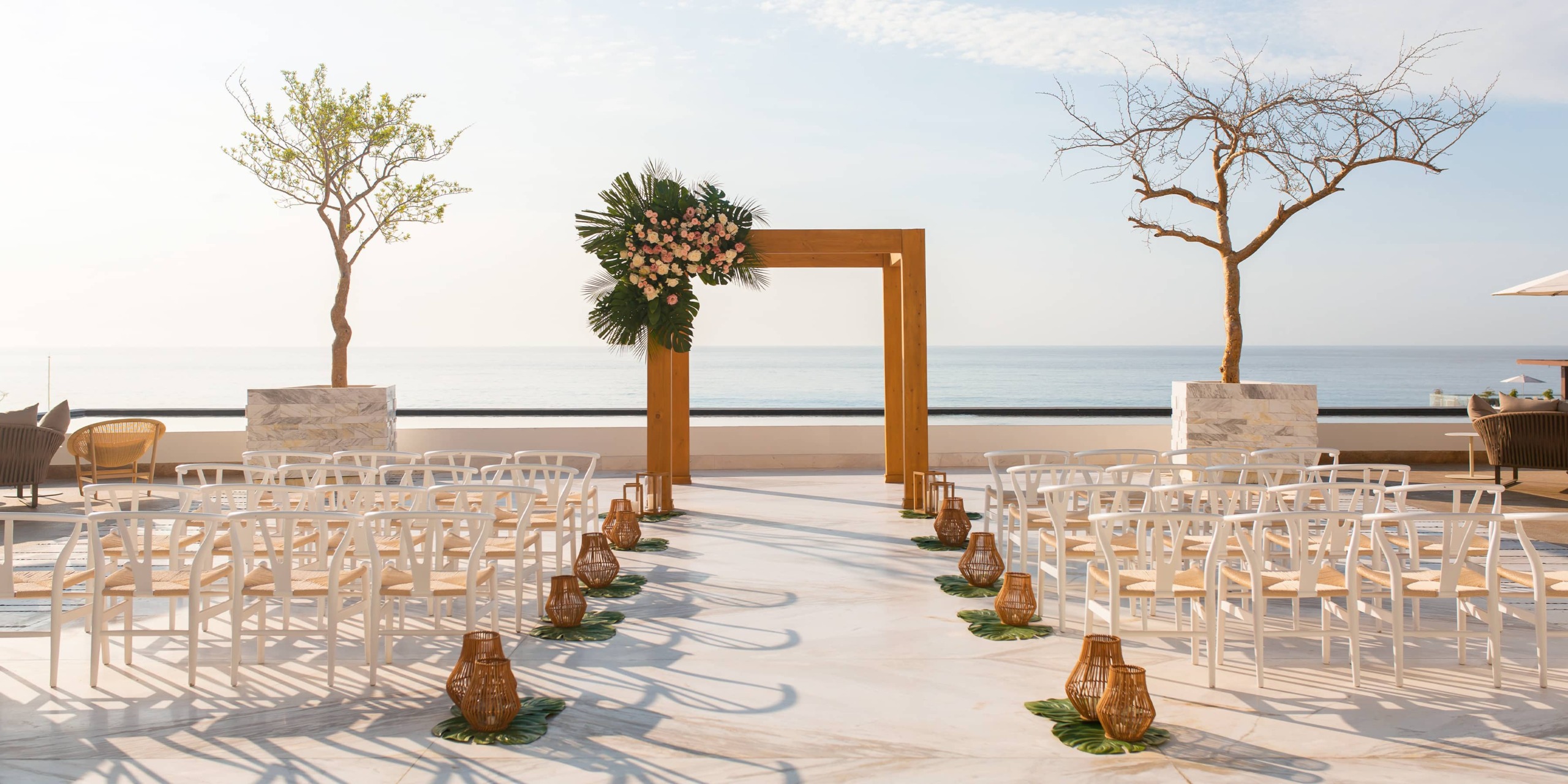 Outdoor wedding ceremony setup by the sea with a Moon Palace The Grand adorned with flowers and surrounded by white chairs arranged in rows. Two trees stand on either side, and lanterns line the aisle.