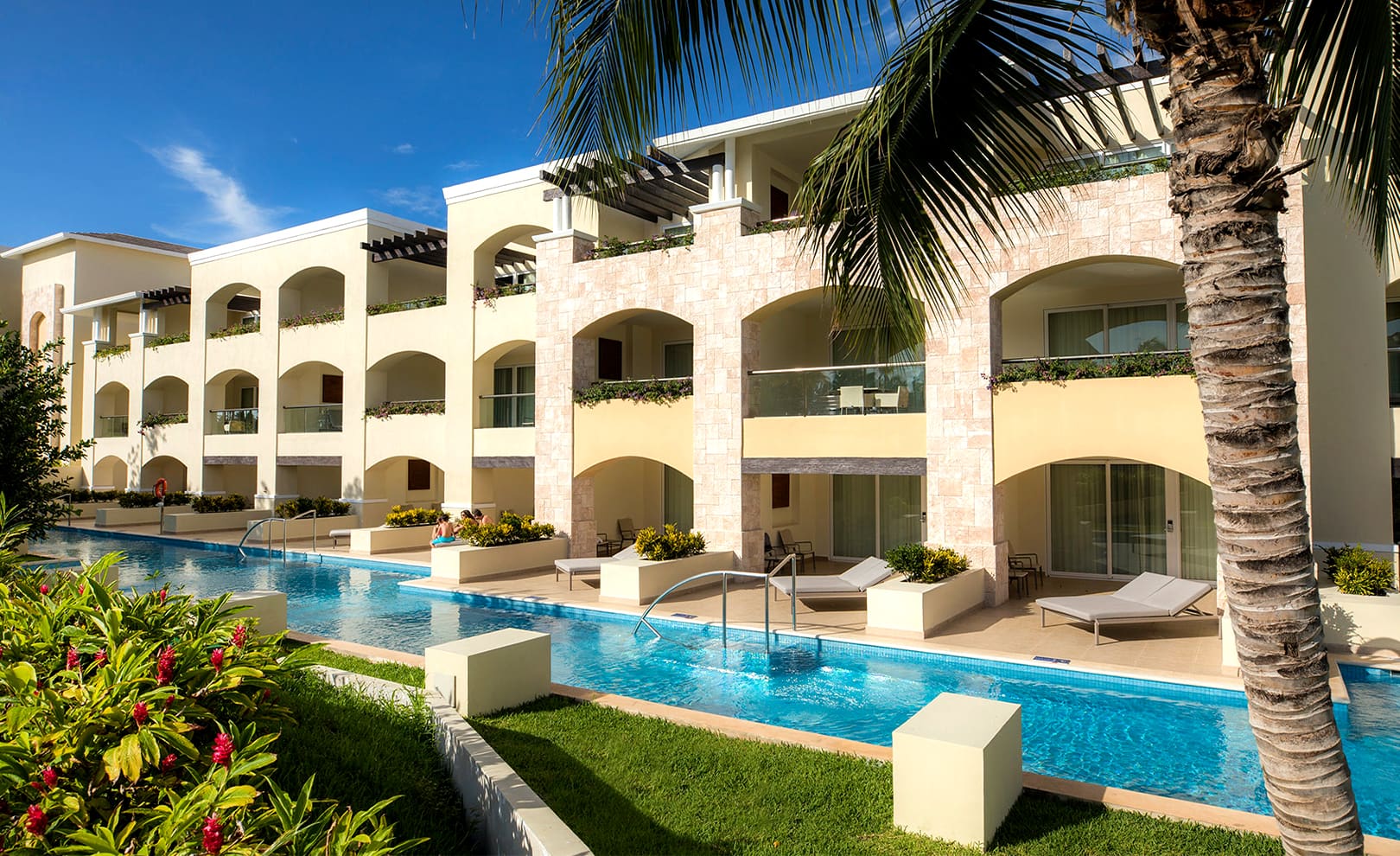 Three-story beige building with balconies and pool access rooms, surrounded by tropical plants and featuring a long, narrow swimming pool in the foreground beneath a clear blue sky at Moon Palace The Grand.