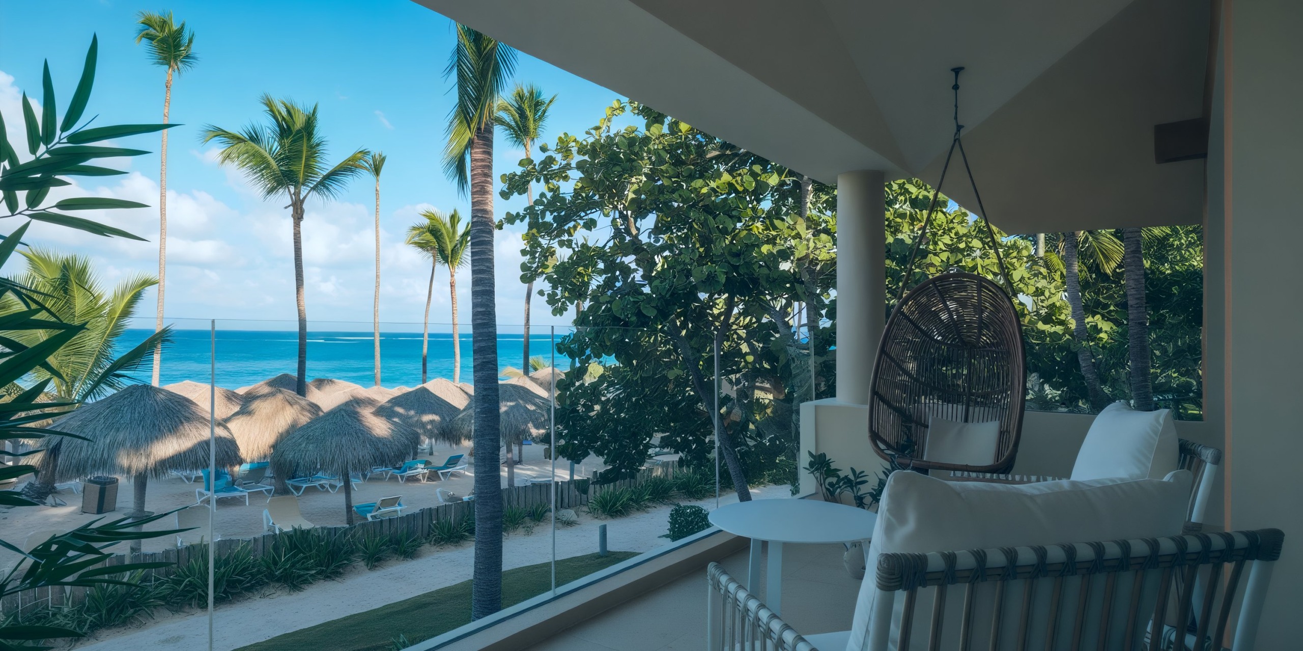 Enjoy the view from your balcony at Coral Level at Iberostar Selection Bavaro, featuring a hanging chair that overlooks a beach adorned with palm trees and thatched umbrellas under a clear blue sky.