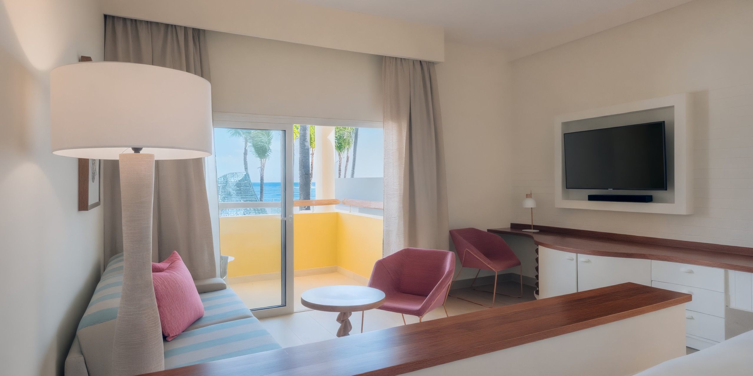 The Coral Level at Iberostar Selection Bavaro offers a modern hotel room with a sofa, two pink chairs, a round table, wall-mounted TV, and a sliding door that opens to a balcony with a pool view.