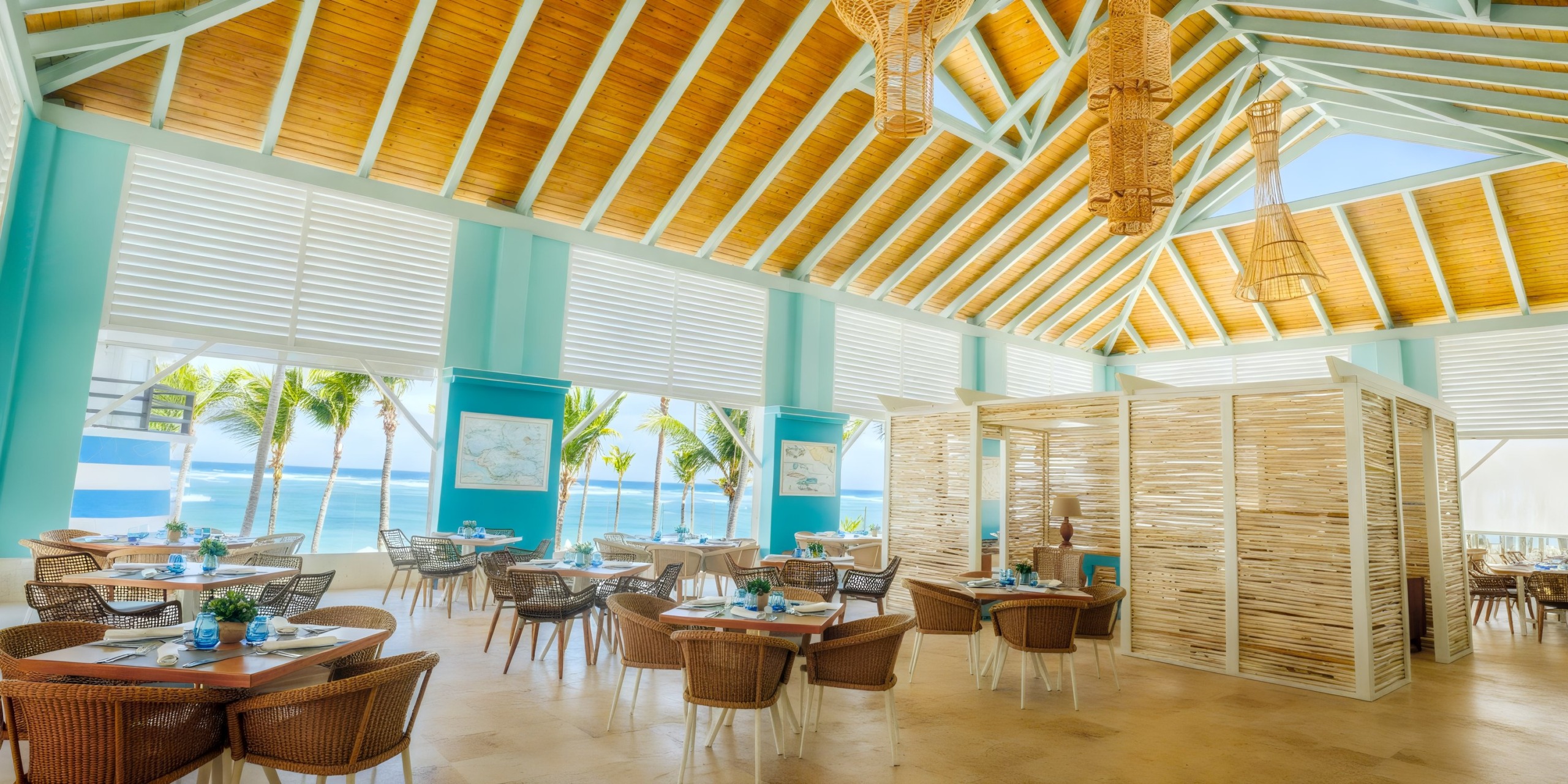 Coral Level at Iberostar Selection Bavaro boasts a bright beachfront setting with wicker chairs, wooden tables, and large windows offering sea views. The high ceiling features woven light fixtures and teal accents, complemented by a stylish partitioned area.