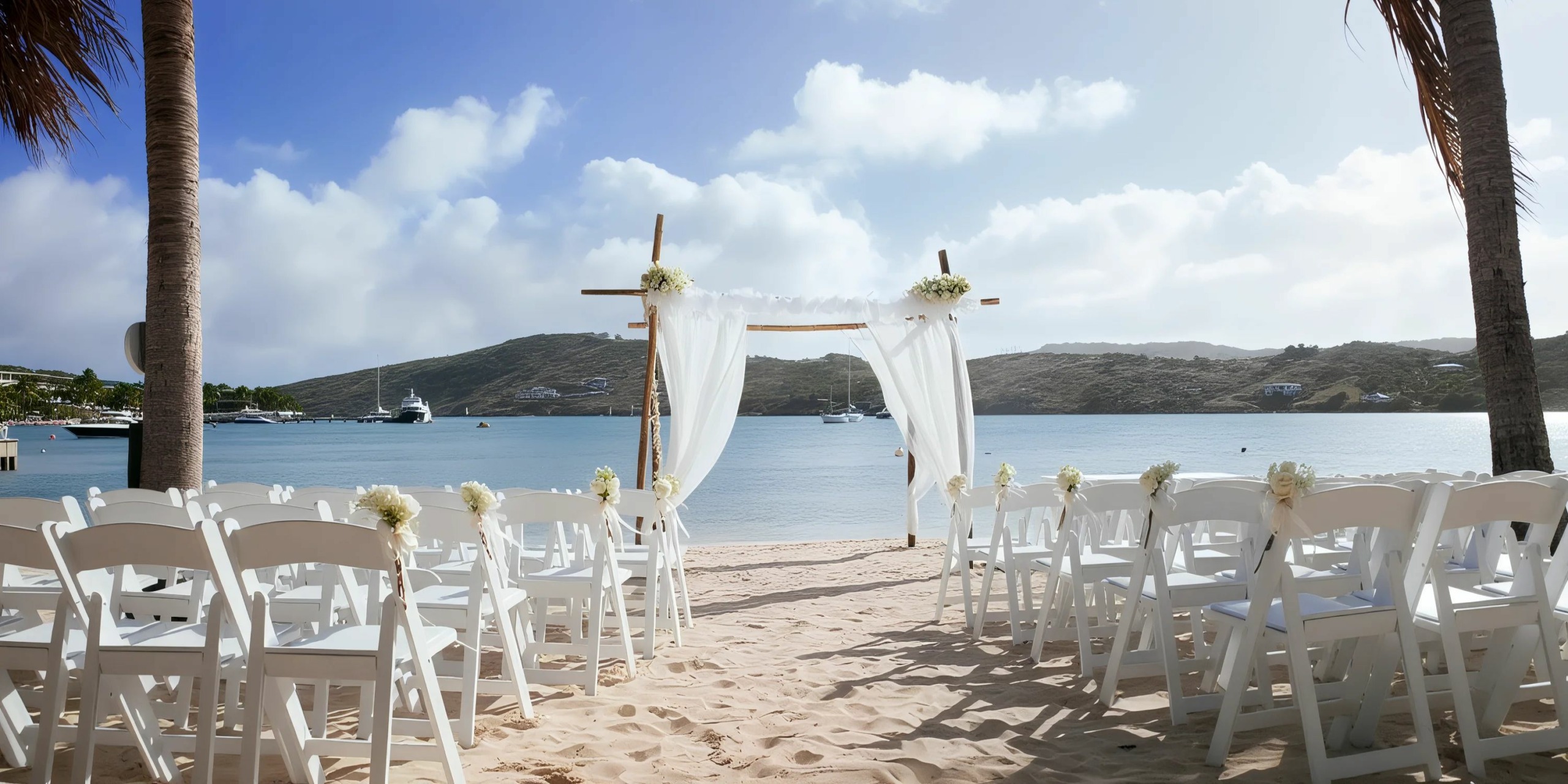 Beach wedding setup with white chairs and an arch draped with white fabric facing a calm sea and distant hills.