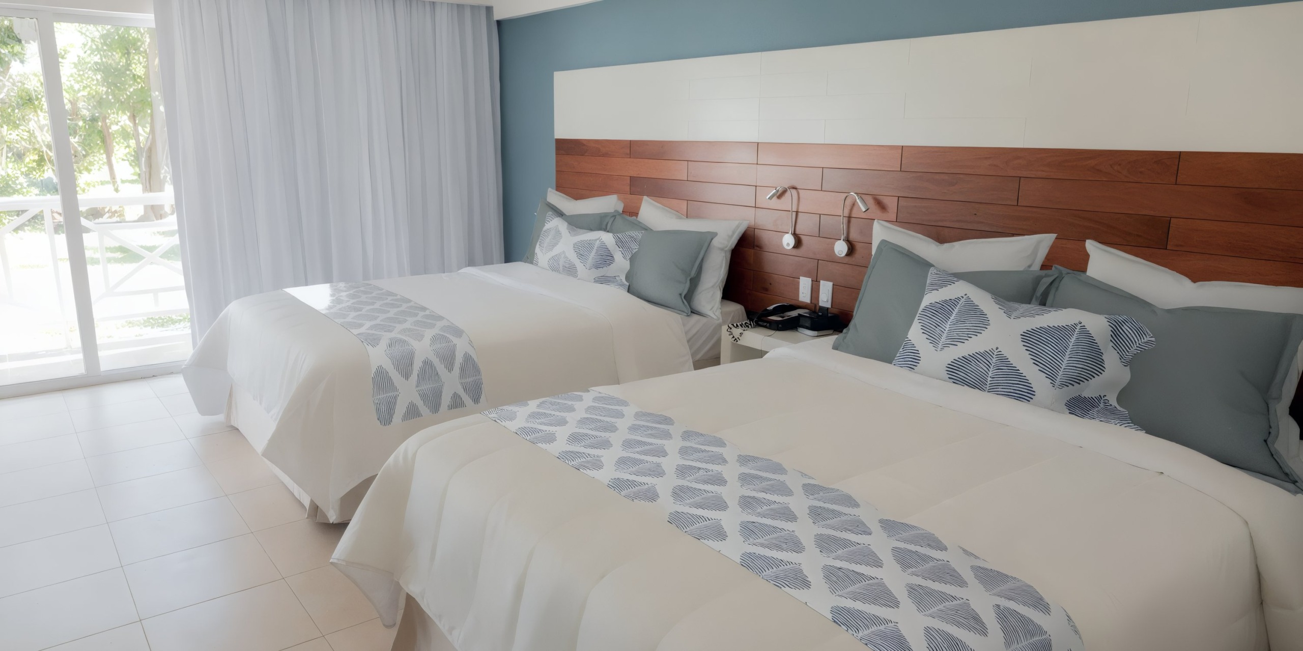 A room at the Viva Wyndham Fortuna Beach features two neatly made double beds with blue and white decor, complemented by a wooden headboard and soft lighting. To the left, there's a window with closed curtains.