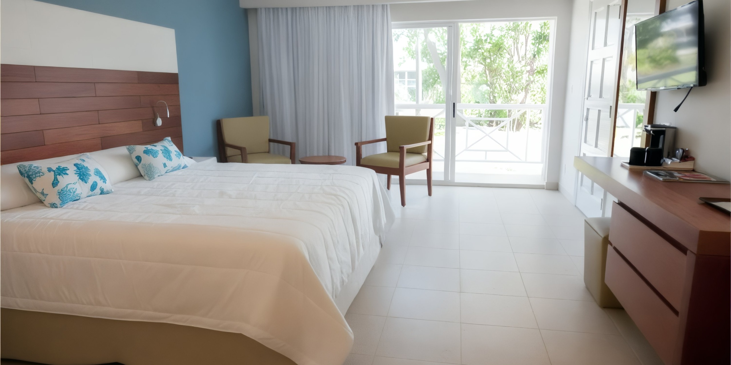 Hotel room at Viva Wyndham Fortuna Beach featuring a spacious bed, two comfortable chairs, a round table, a wall-mounted TV, and a desk. A bright window adorned with sheer curtains allows for ample natural light.