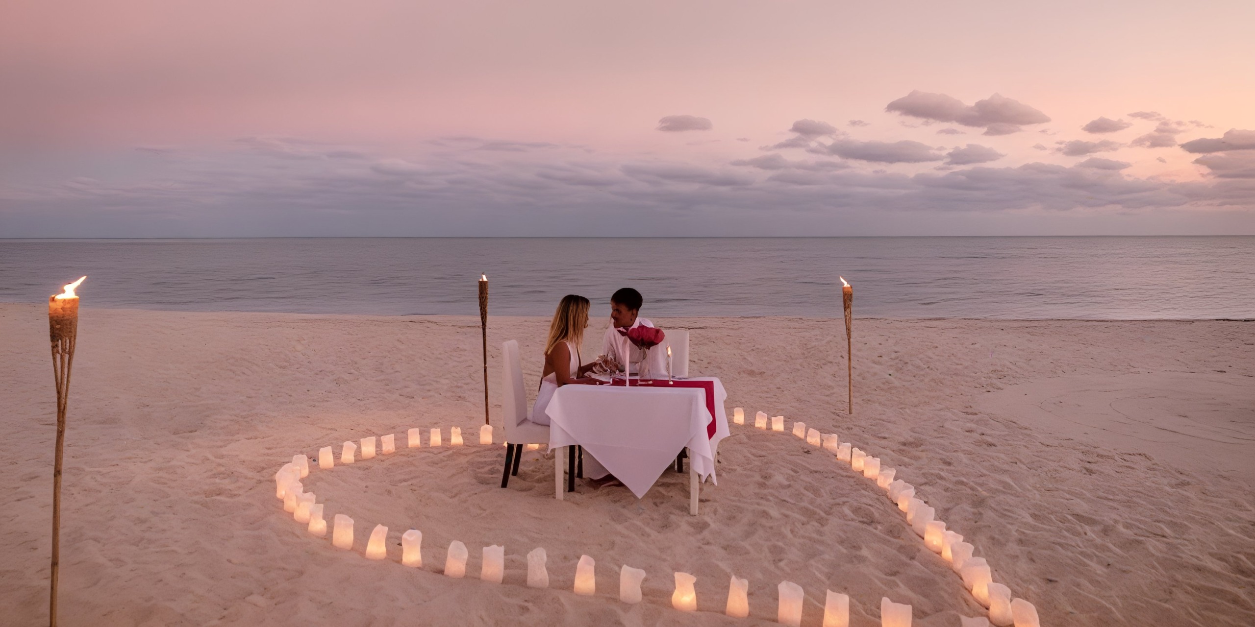 A couple enjoys a romantic dinner at a candlelit table on the beach, framed by a heart-shaped arrangement of candles, with the ocean providing a stunning backdrop beneath the cloudy evening sky at Viva Wyndham Fortuna Beach.