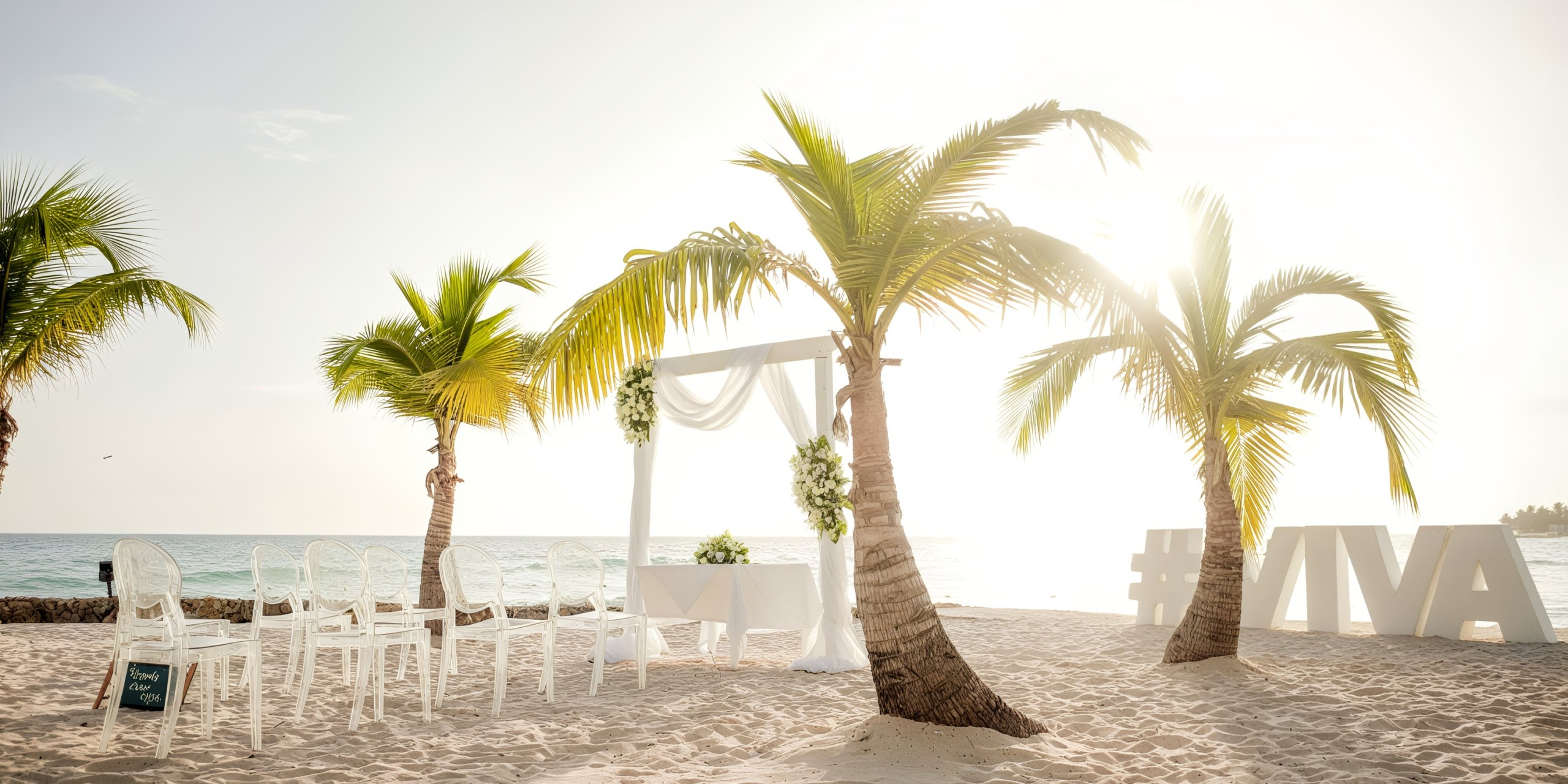 A beach wedding setup at Viva Wyndham Fortuna Beach features white chairs, a floral arch, and palms on the sandy shore. Large "#VIVA" letters enhance the picturesque ocean view in the background.