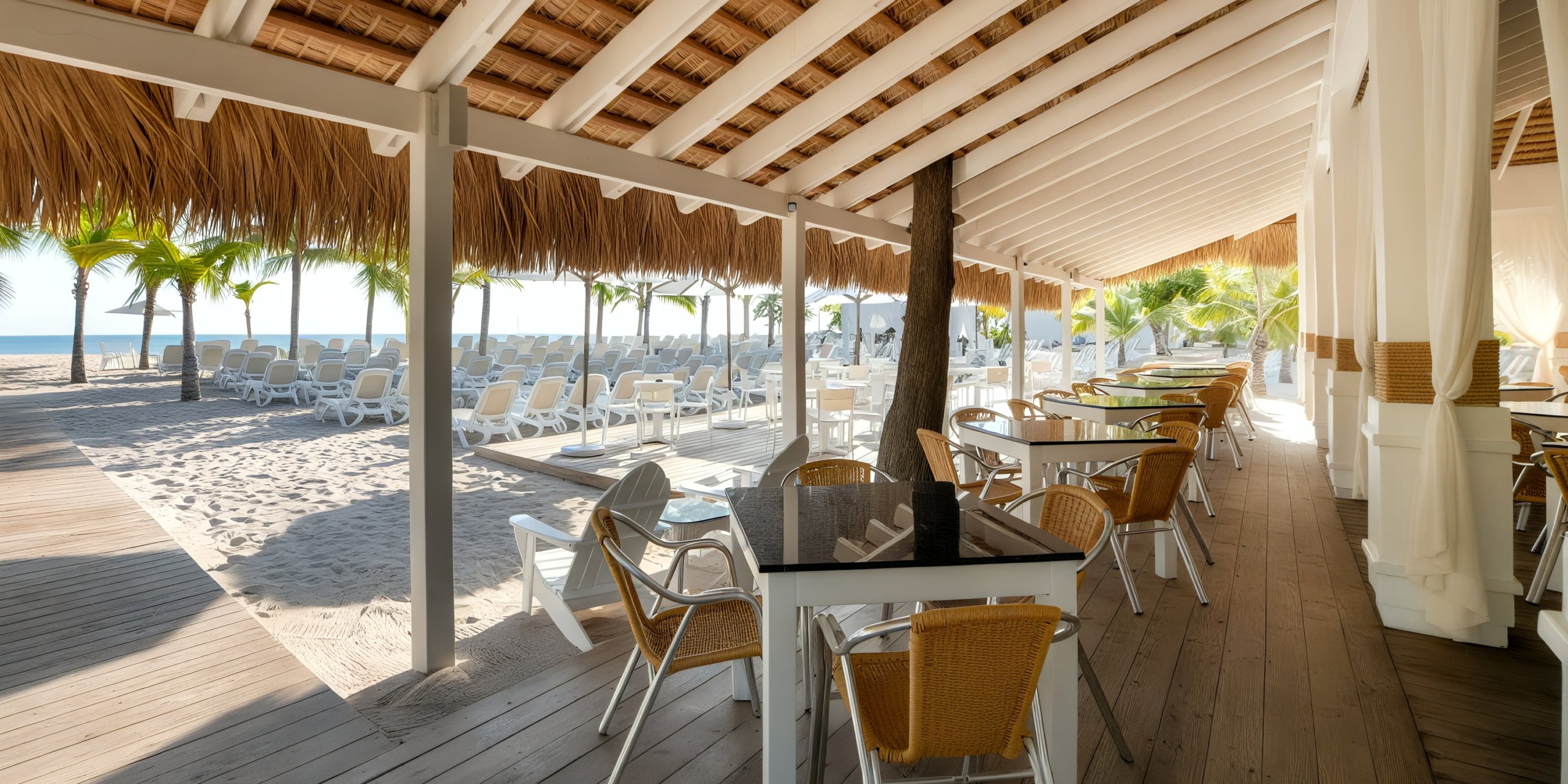 Enjoy dining at Viva Wyndham V Heavens - All-Inclusive Resort, Adults Only, featuring a charming outdoor beachside restaurant with wicker chairs and tables nestled under a thatched roof. Relax on the lounge chairs while taking in the views of palm trees lining the sandy beach in the background.