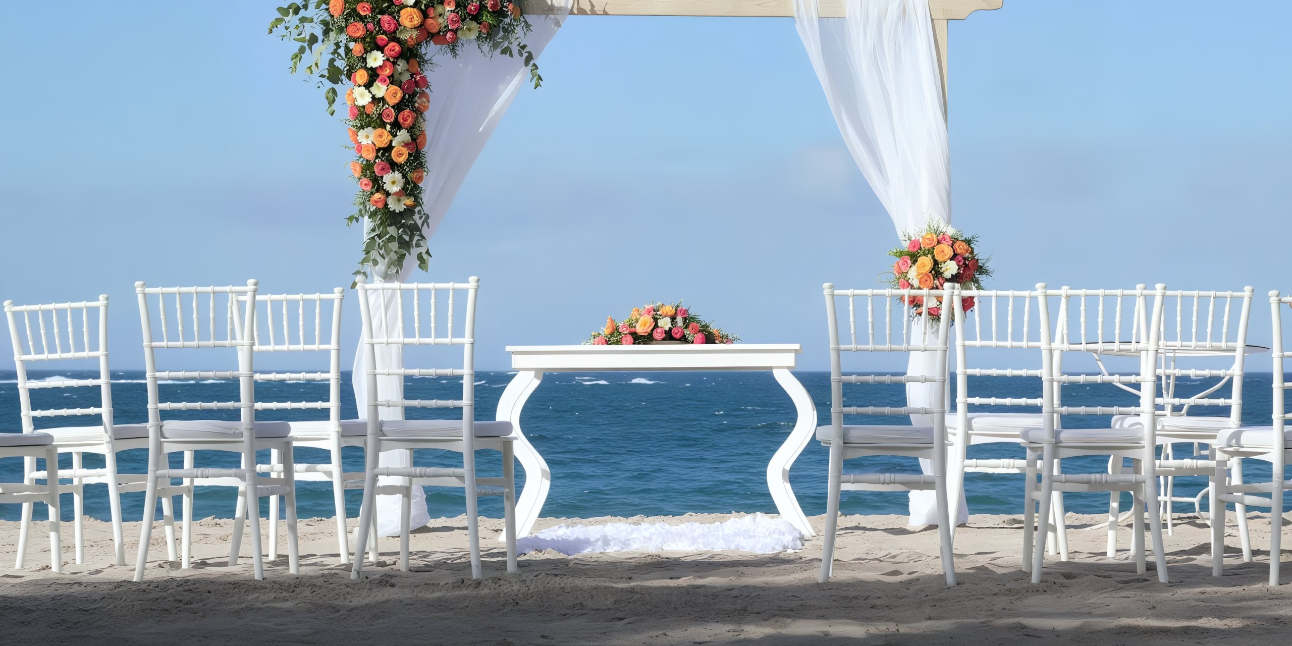 The beach wedding setup at Viva Wyndham V Heavens - All-Inclusive Resort, Adults Only features a white arch beautifully decorated with flowers and sheer drapes. White chairs are arranged facing a table enhanced with floral arrangements, set against the stunning backdrop of the ocean.