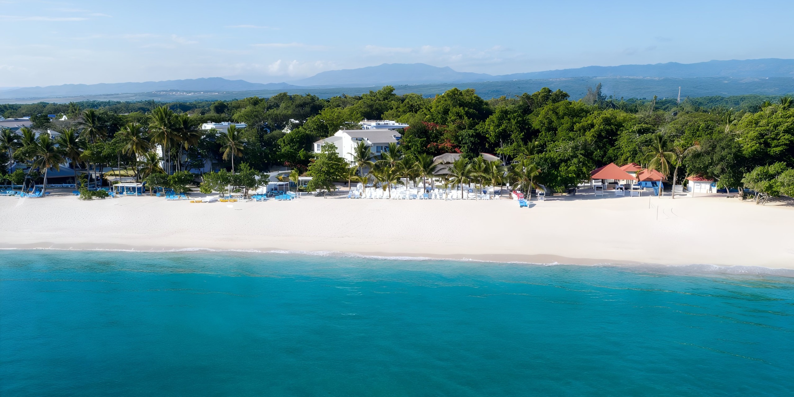 Aerial view of the Viva Wyndham V Heavens - All-Inclusive Resort, Adults Only, nestled amidst white sandy beaches and crystal-clear turquoise waters. The resort is surrounded by lush green trees with buildings subtly integrated into the natural landscape. Majestic mountains create a stunning backdrop in the distance.