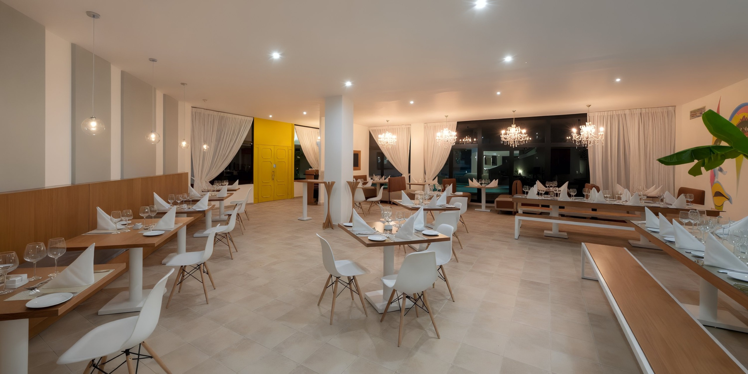 Step into Viva Wyndham V Heavens - All-Inclusive Resort, Adults Only, where the modern restaurant features stylish wooden tables paired with sleek white chairs. Large windows draped with elegant white curtains and well-placed ceiling lights contribute to a bright and inviting atmosphere.