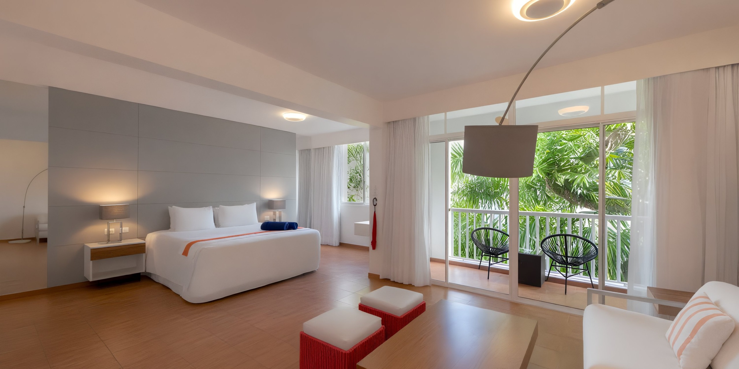 A contemporary room at Viva Wyndham V Heavens - All-Inclusive Resort, Adults Only, featuring a large bed, side tables with lamps, a sitting area, and a balcony with views of lush greenery.
