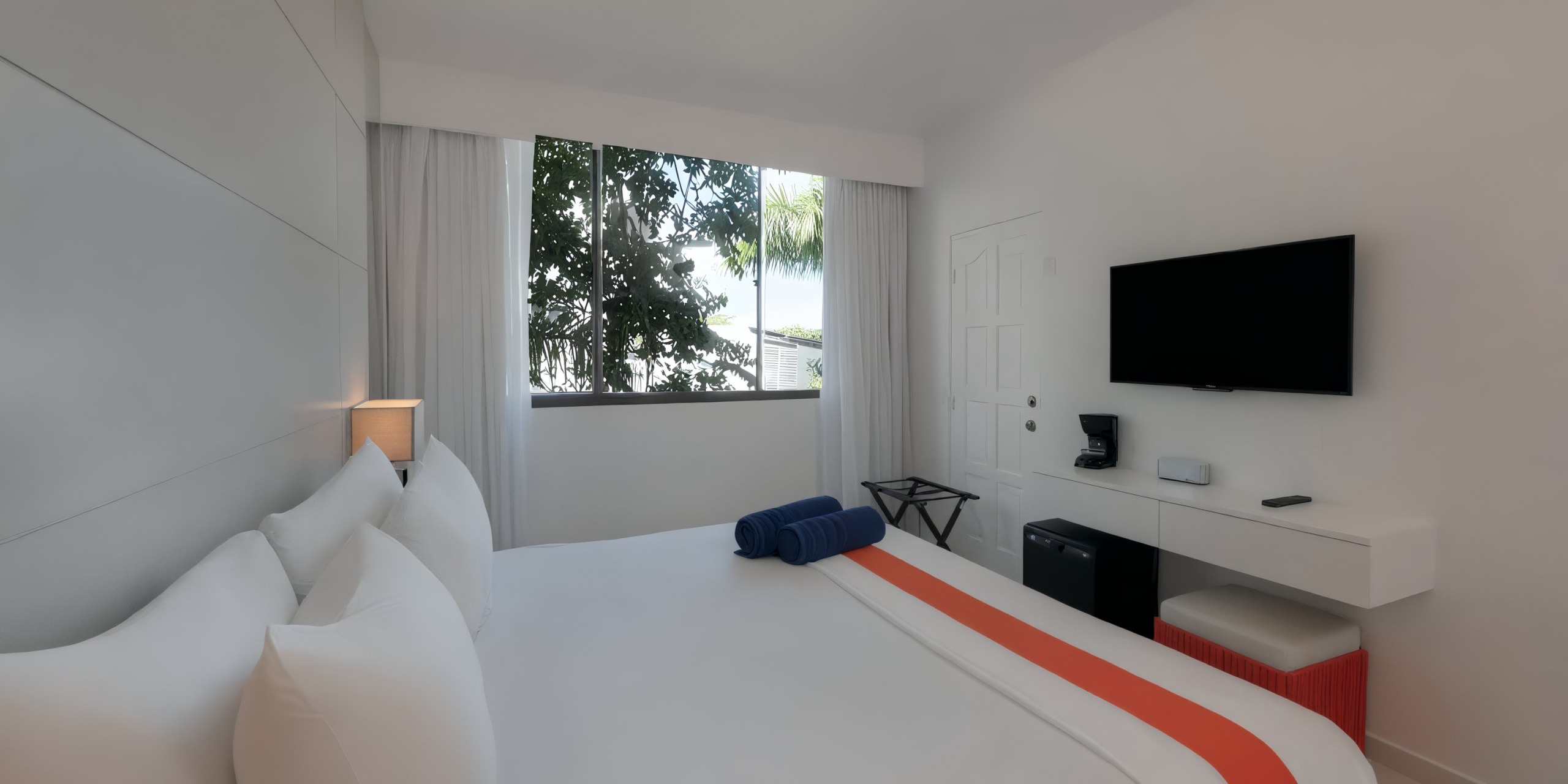 Experience the Viva Wyndham V Heavens - All-Inclusive Resort, Adults Only, where the minimalist hotel room features a spacious bed adorned with white and orange bedding. The room includes a wall-mounted TV and offers a garden view through its window. Enjoy the convenience of a small desk area complete with a coffee maker for your comfort.