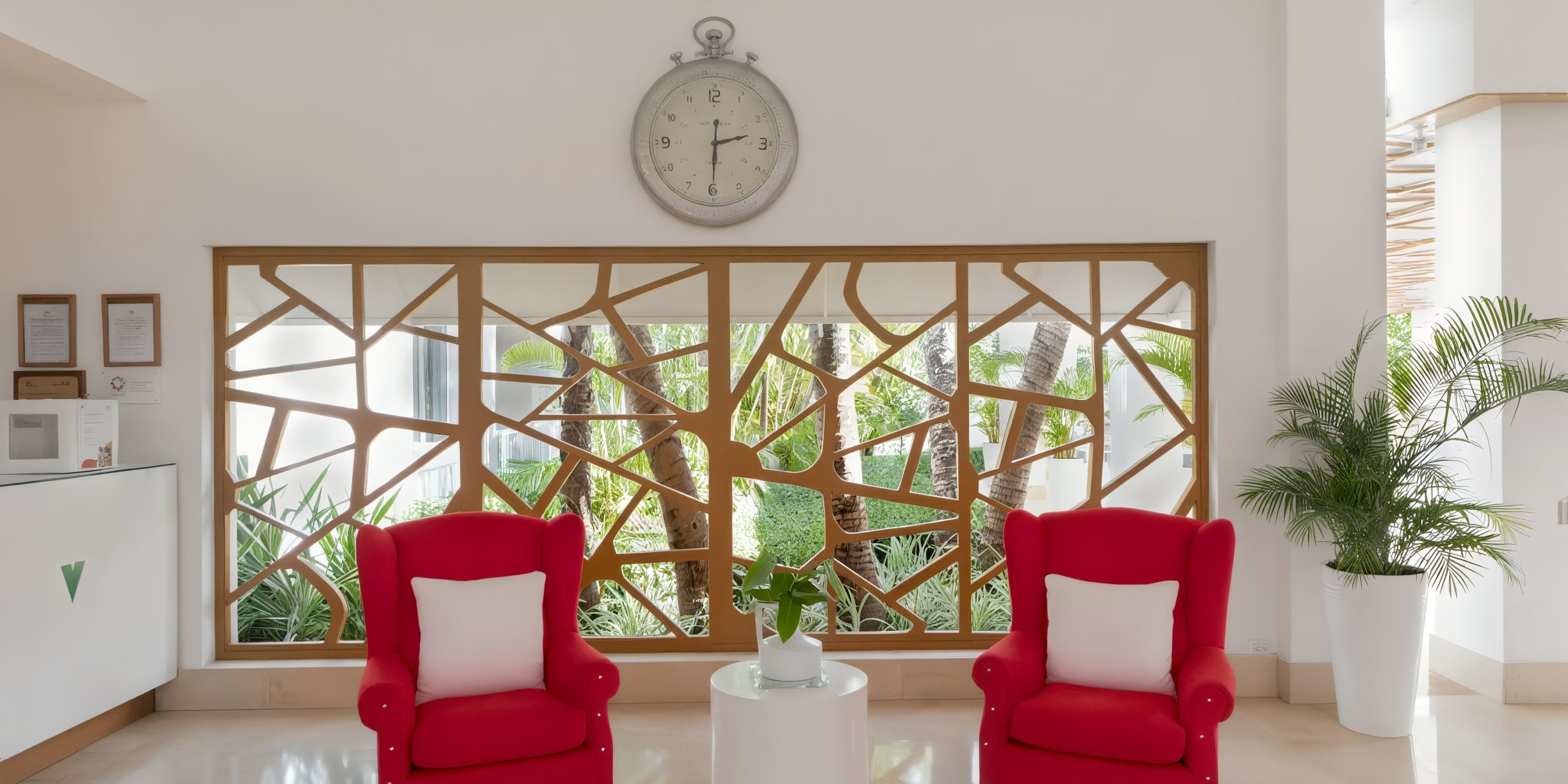 In the waiting area of the Viva Wyndham V Heavens - All-Inclusive Resort, Adults Only, you'll find two red armchairs paired with a white round table and a decorative partition in the background. A large clock adorns the wall above.