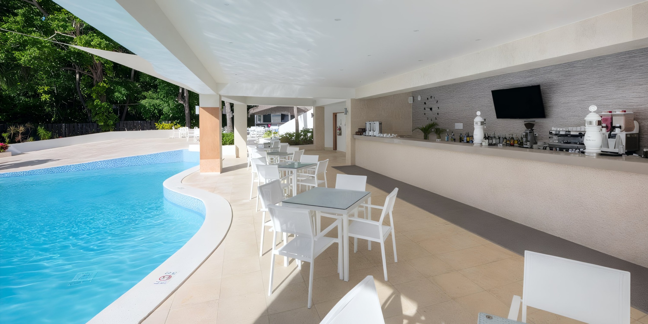 At the Viva Wyndham V Heavens - All-Inclusive Resort, Adults Only, you’ll find a poolside bar area equipped with white tables and chairs alongside a counter stocked with drinks and a television. The pool is conveniently situated to the left, while lush greenery serves as the backdrop.
