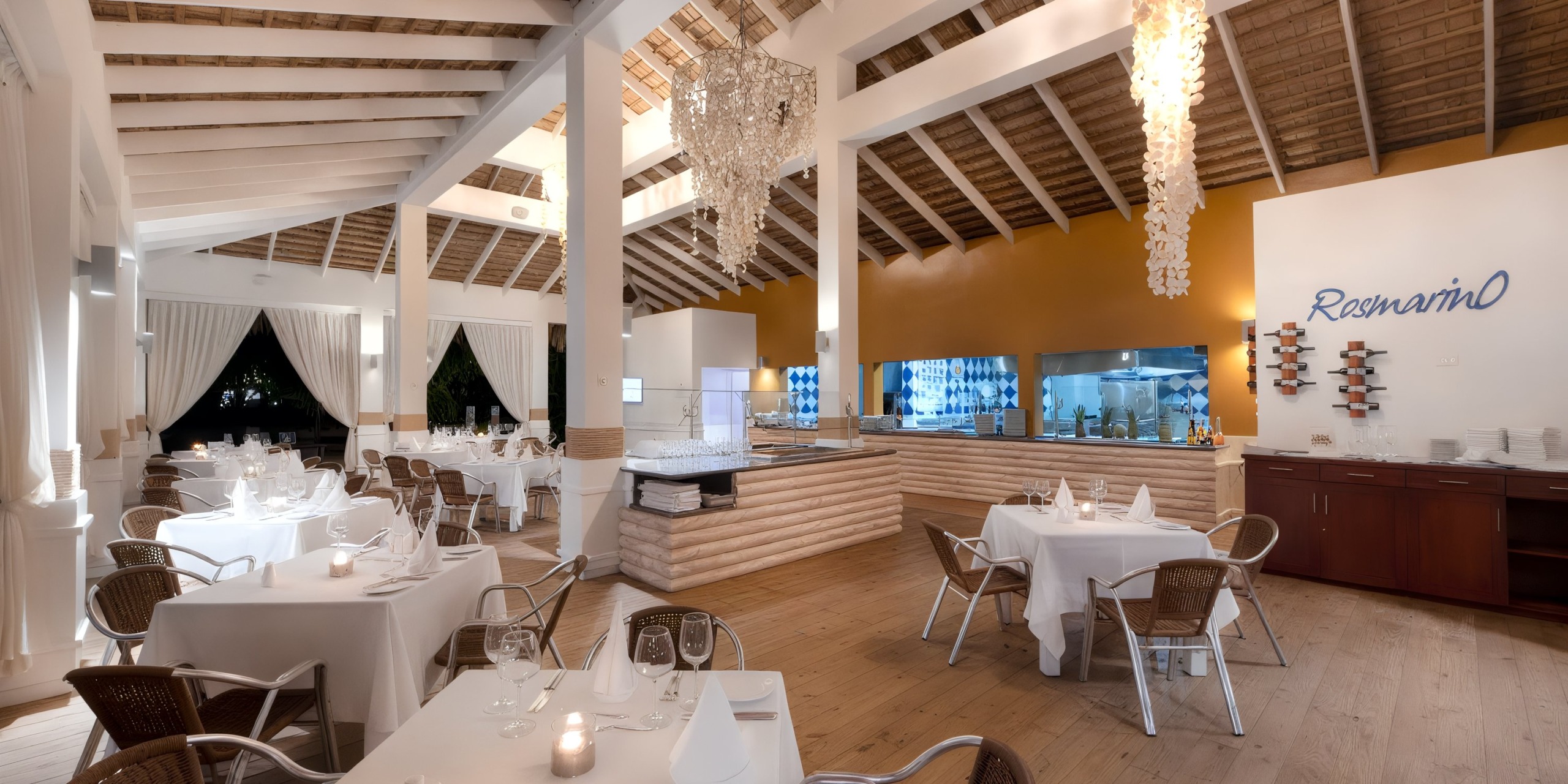 The sophisticated interior of the Viva Wyndham V Heavens - All-Inclusive Resort, Adults Only features white tablecloths, wicker chairs, and chandeliers. In the background, you can see the open kitchen and a sign that reads "Rosmarino" on the wall.
