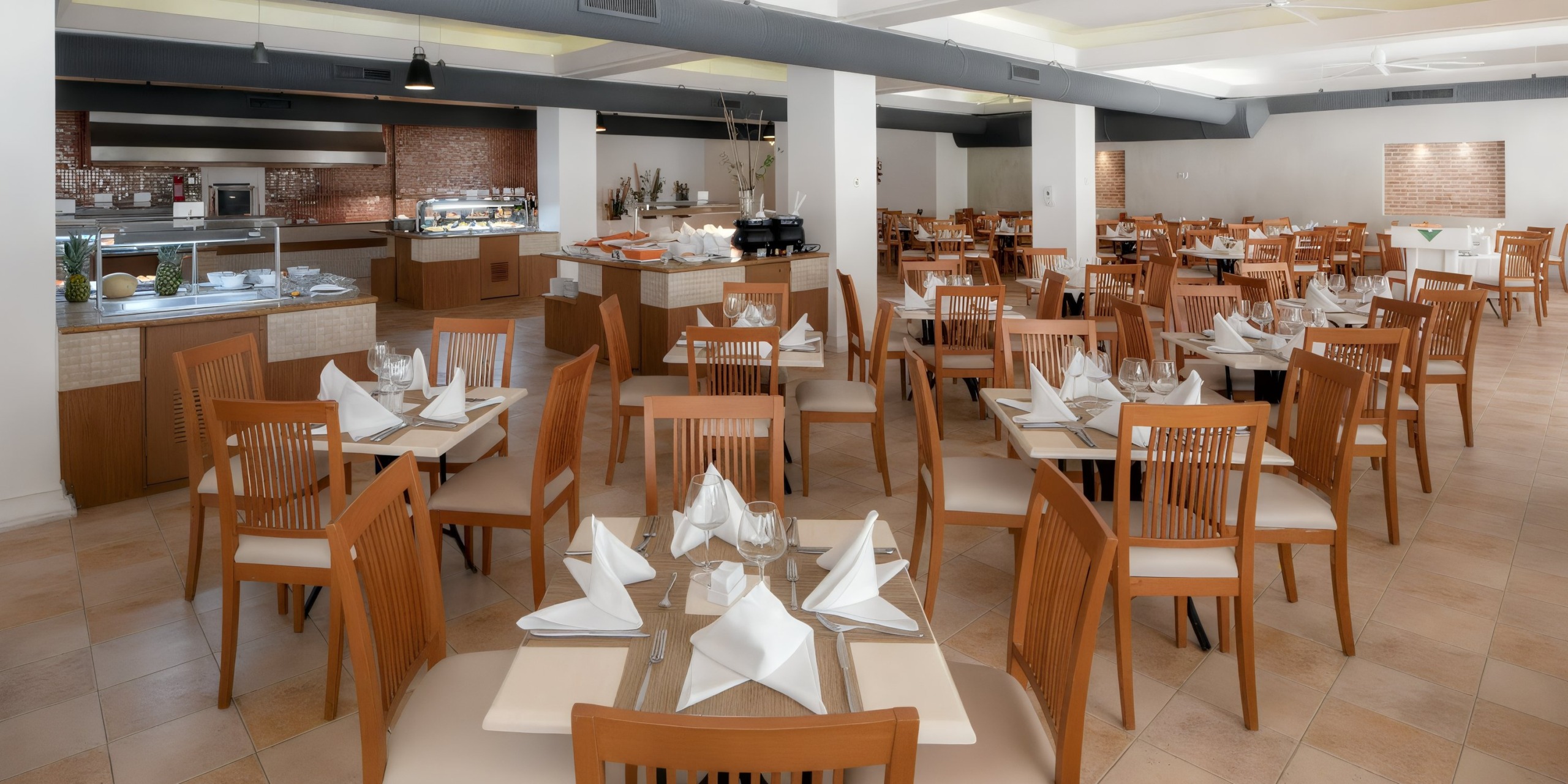 The Viva Wyndham V Heavens - All-Inclusive Resort, Adults Only features a spacious dining area with neatly arranged wooden tables and chairs, each beautifully set with folded napkins and tableware. In the background, guests can catch a glimpse of the inviting buffet area.